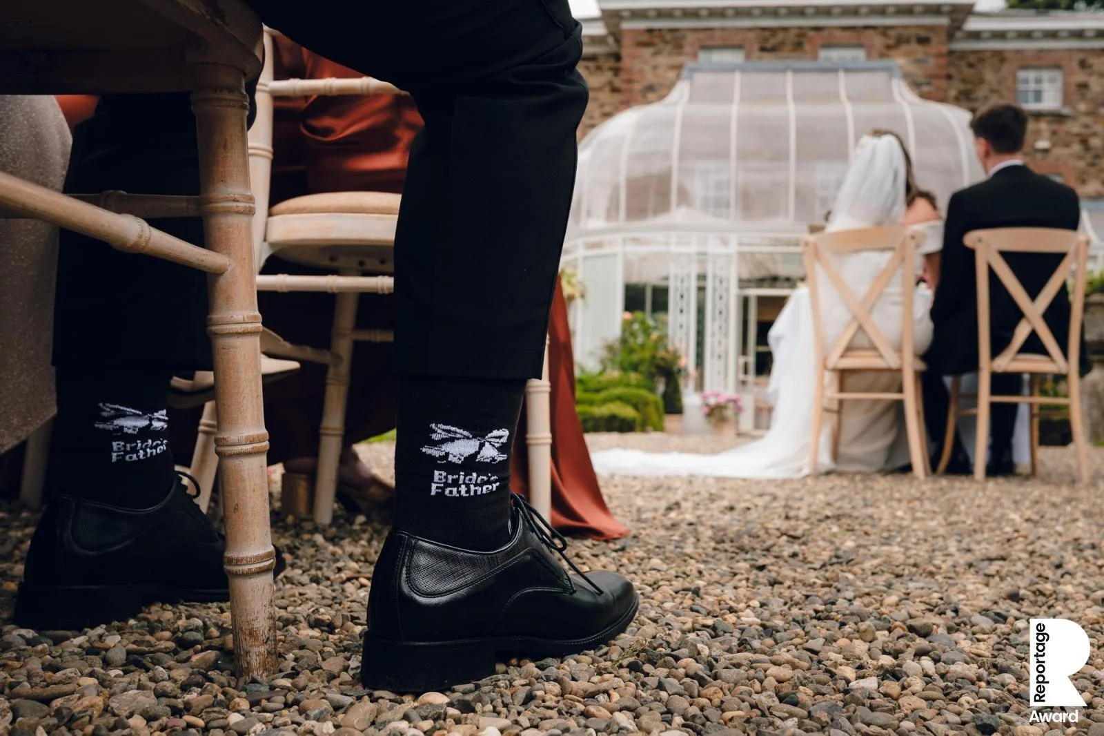 Close-up of a man wearing black dress shoes, black socks with wedding-themed text and graphics, sitting at a wedding reception. In the background, a bride and groom sit at a table outdoors, with a glass and metal structure behind them.