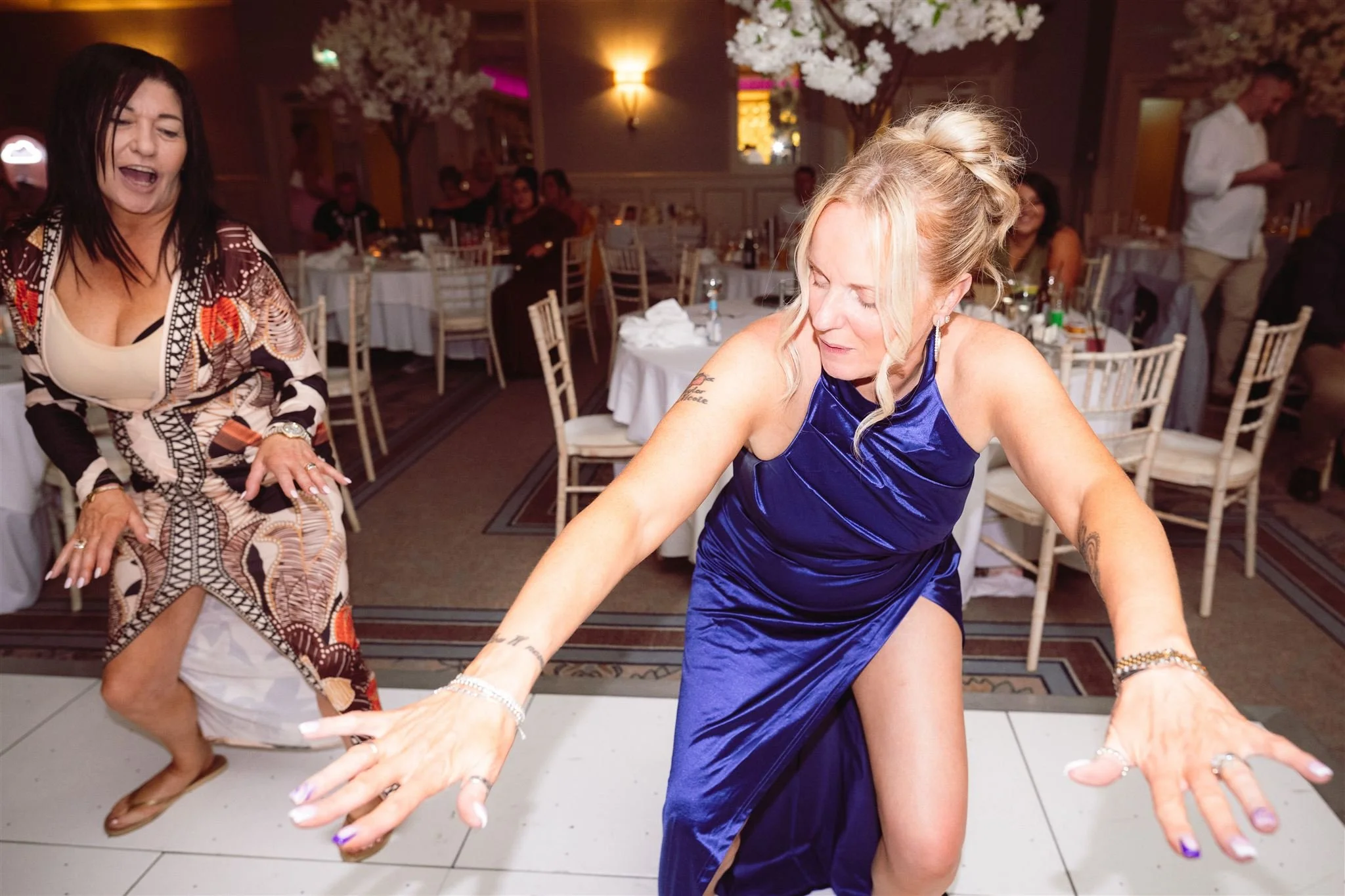 Two women dancing at an indoor event with decorated tables and floral centerpieces.