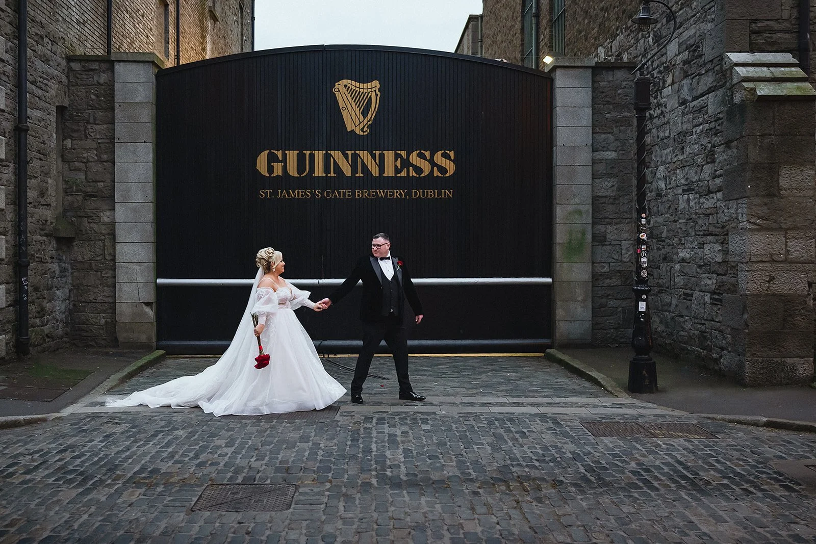 A bride and groom holding hands in front of a large black Guinness brewery gate in Dublin. The bride is in a white wedding dress holding a bouquet of red roses, and the groom is in a black tuxedo with a bow tie.