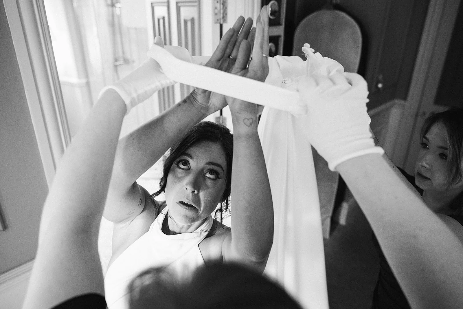 A woman in a white dress, likely a bride, is looking up while her arms are raised, as her wedding gown is being helped or adjusted by another woman wearing white gloves. The scene is in black and white, with a focus on the bride's expressive face and