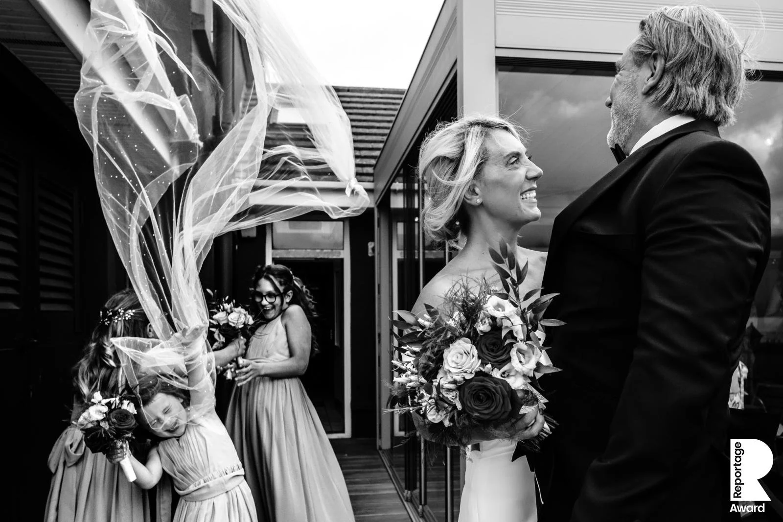 Black and white photograph of a bride and groom smiling and looking at each other on their wedding day, with bridesmaids celebrating in the background.