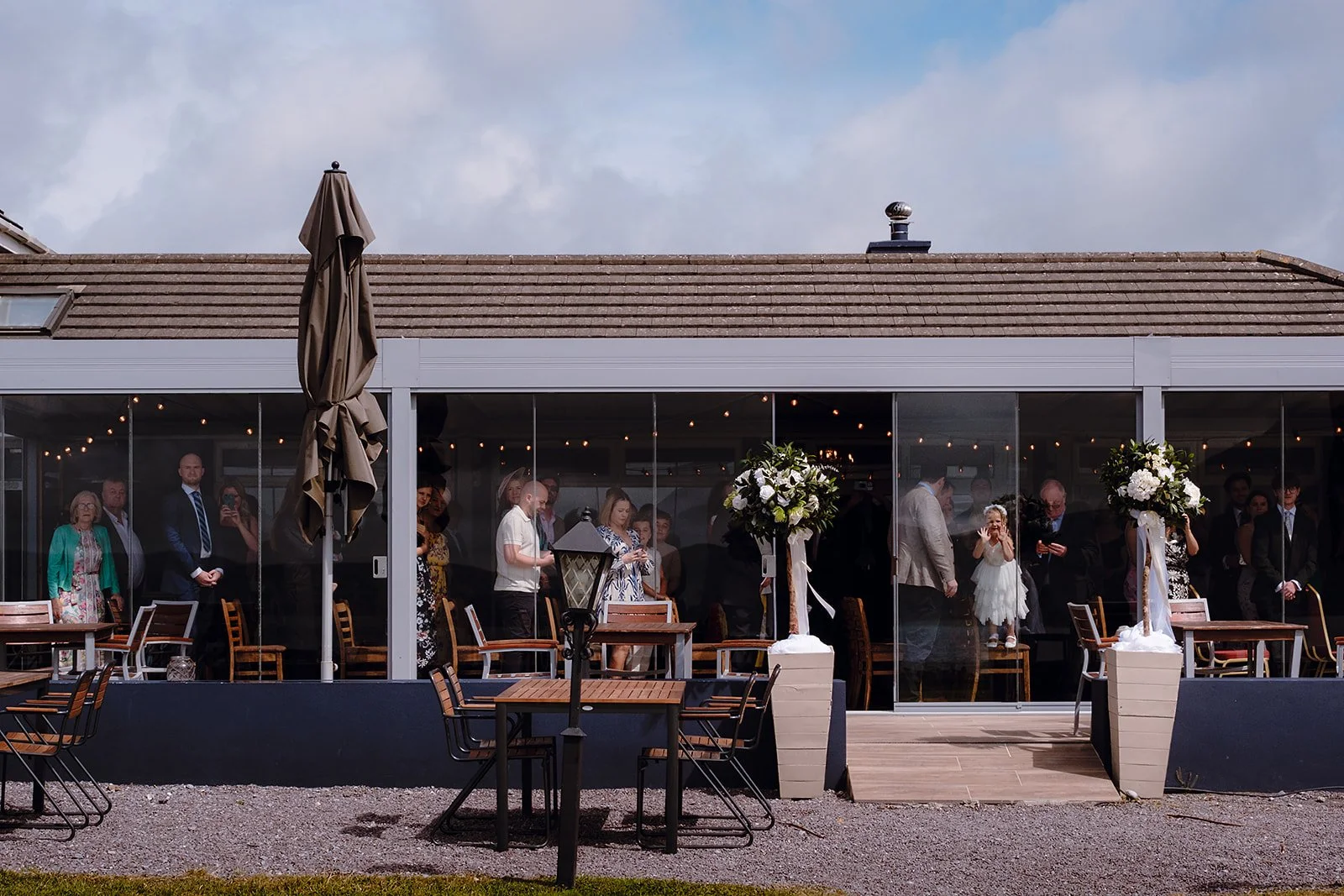 People gathered inside a glass-enclosed venue at a wedding reception, visible through the glass walls, with floral decorations and chairs outside on a deck.