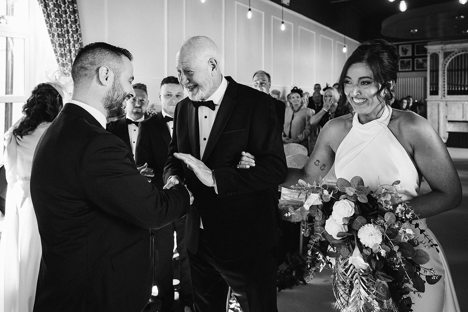 A black-and-white photo of a wedding ceremony. The groom is shaking hands with an older man in a tuxedo who is smiling. The bride, holding a bouquet of flowers, is smiling and watching the handshake. Guests are in the background, observing the event.