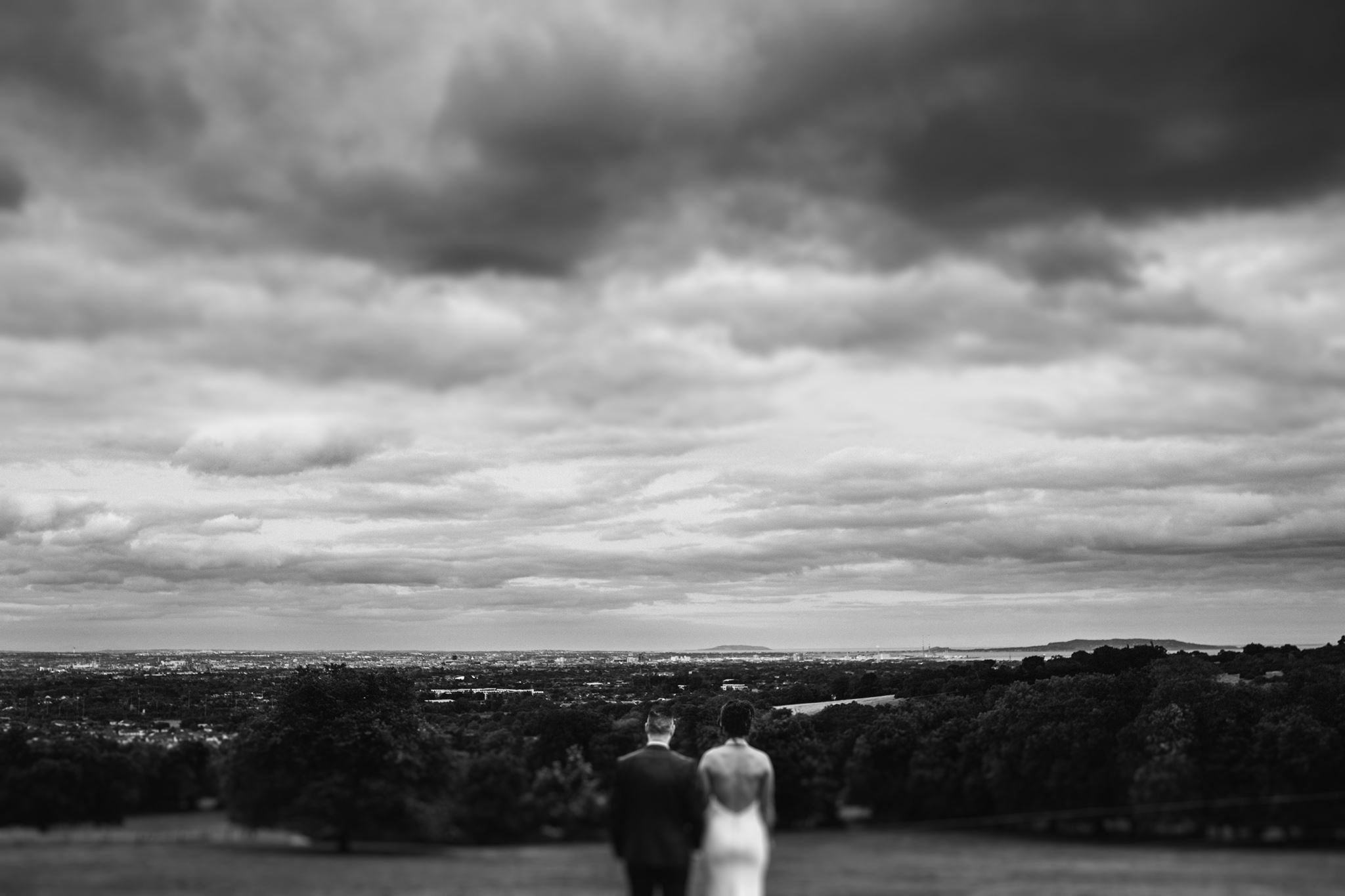 Two men in suits walking on a path in an outdoor area with a cityscape and a cloudy sky in the background.