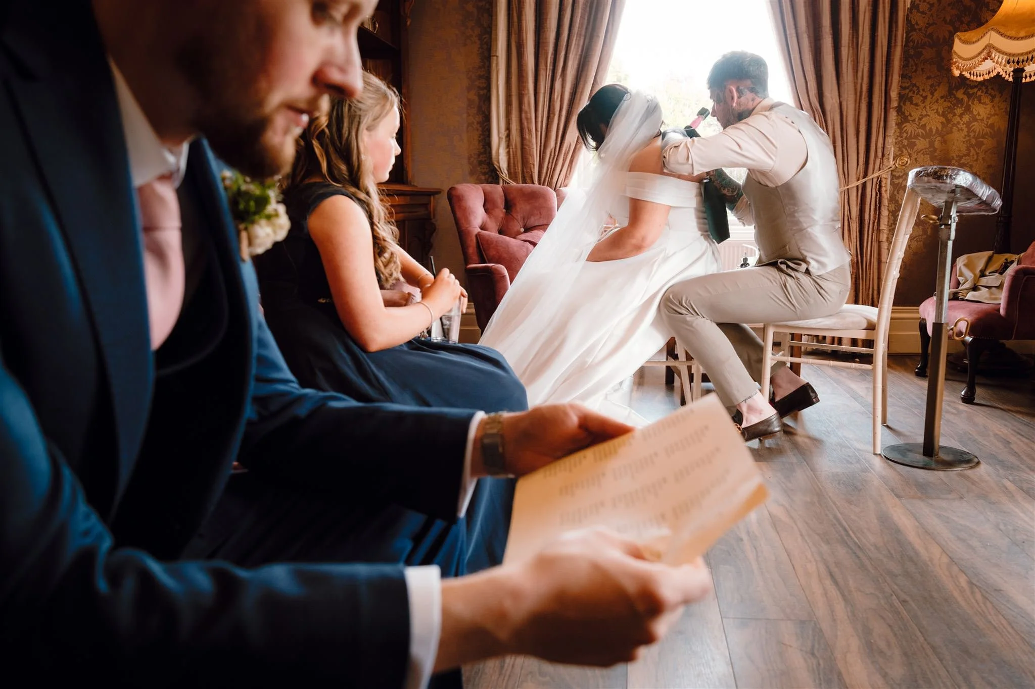 Bride and groom during their wedding ceremony, kneeling on chairs with officiant holding a microphone; guests seated nearby, reading from papers, in a warmly decorated room with curtains and vintage furniture.
