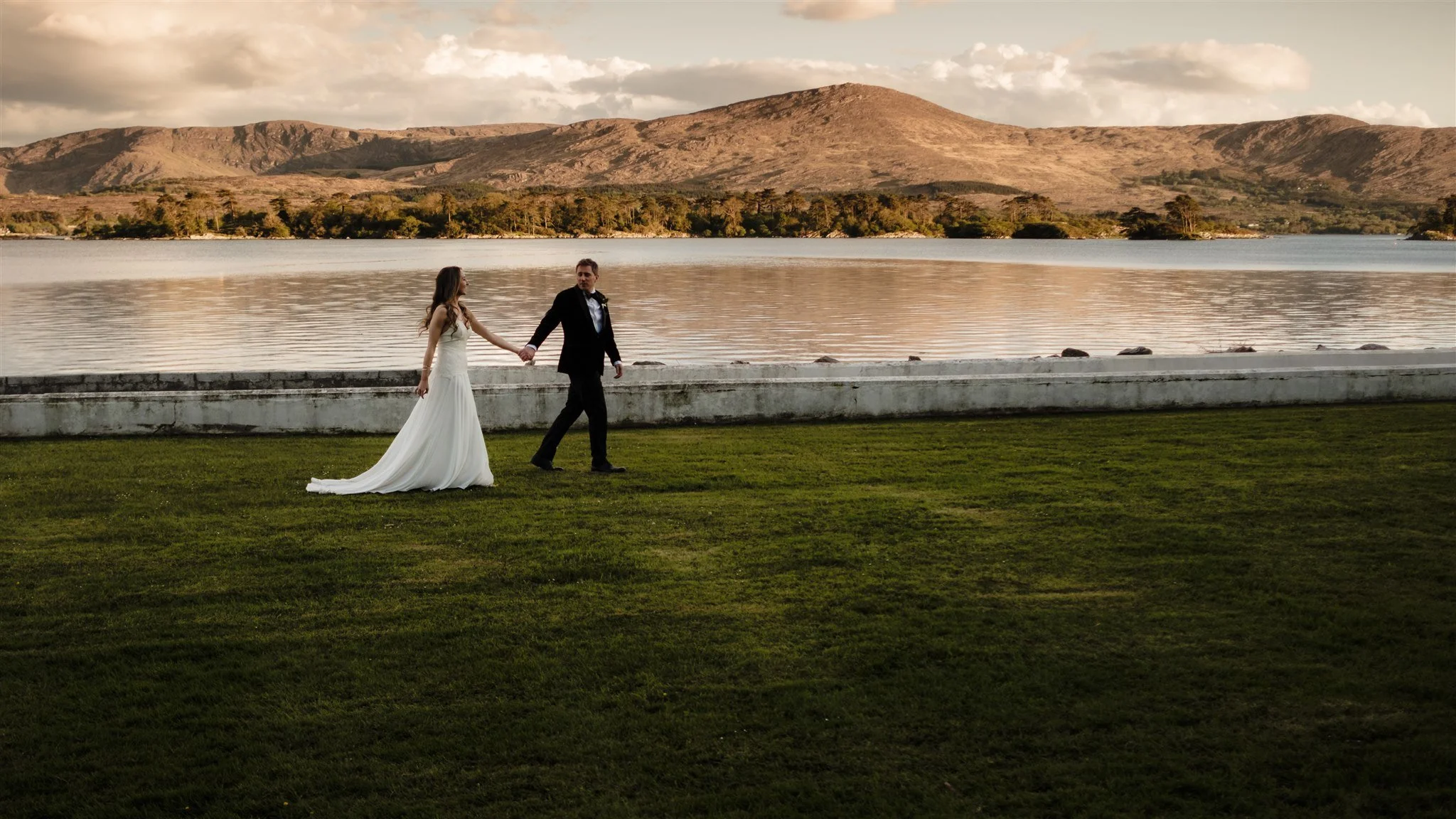 A bride and groom holding hands walking near a body of water with mountains in the background during sunset.