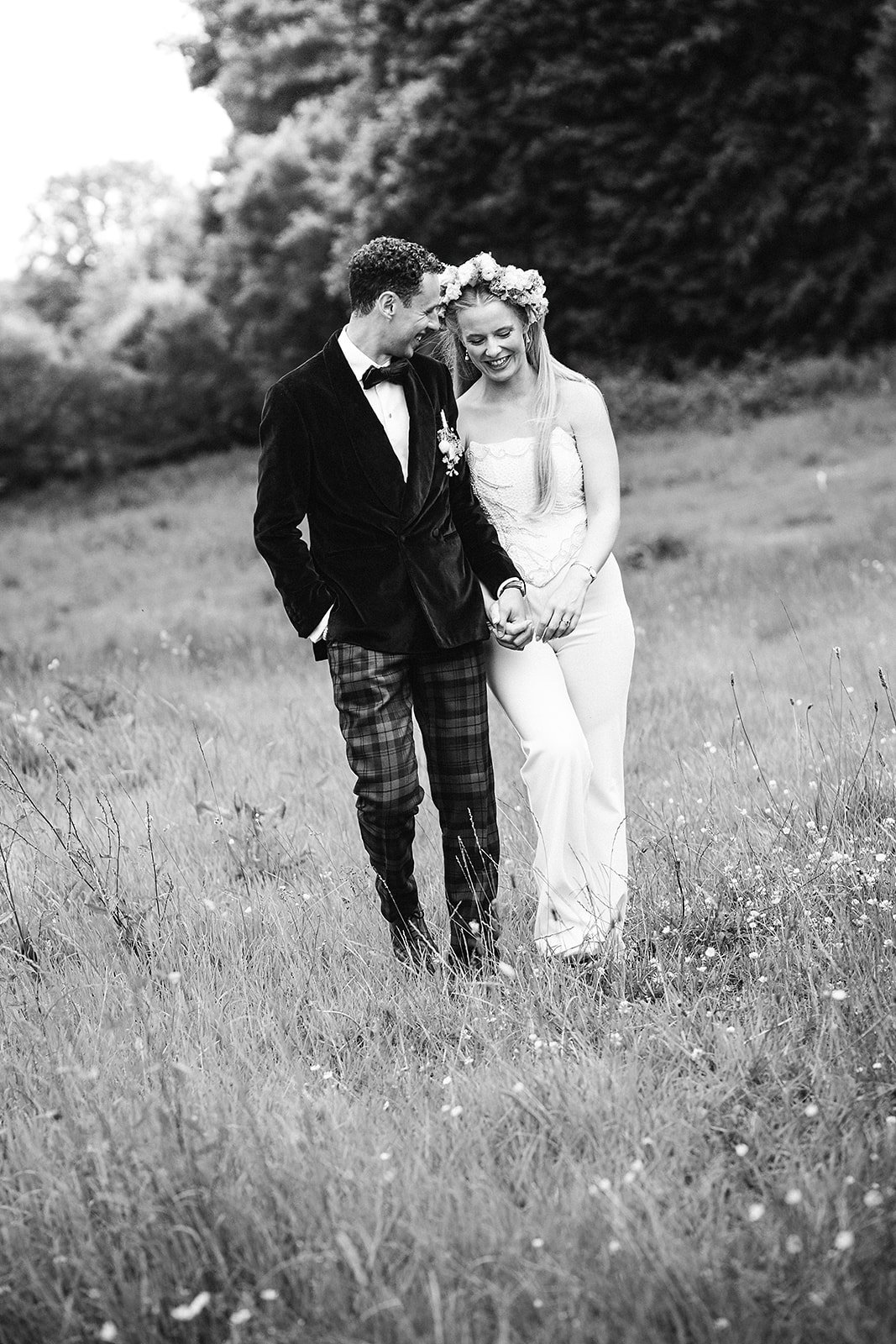 A black and white photo of a smiling couple walking hand in hand through a grassy field, dressed in formal attire, with trees in the background.