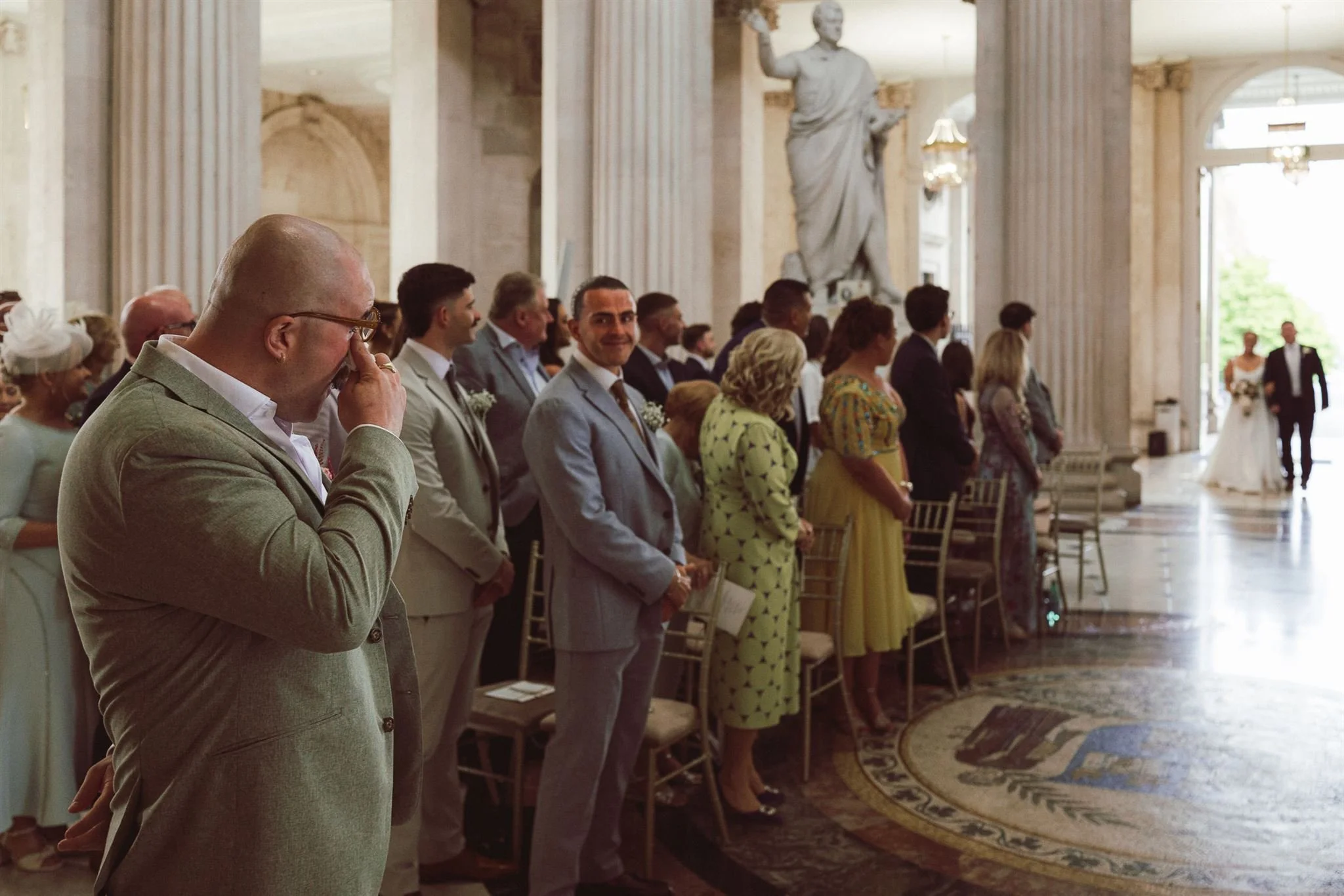 People dressed in formal attire standing in a row during a wedding ceremony inside a grand hall with columns and a large statue in the background.