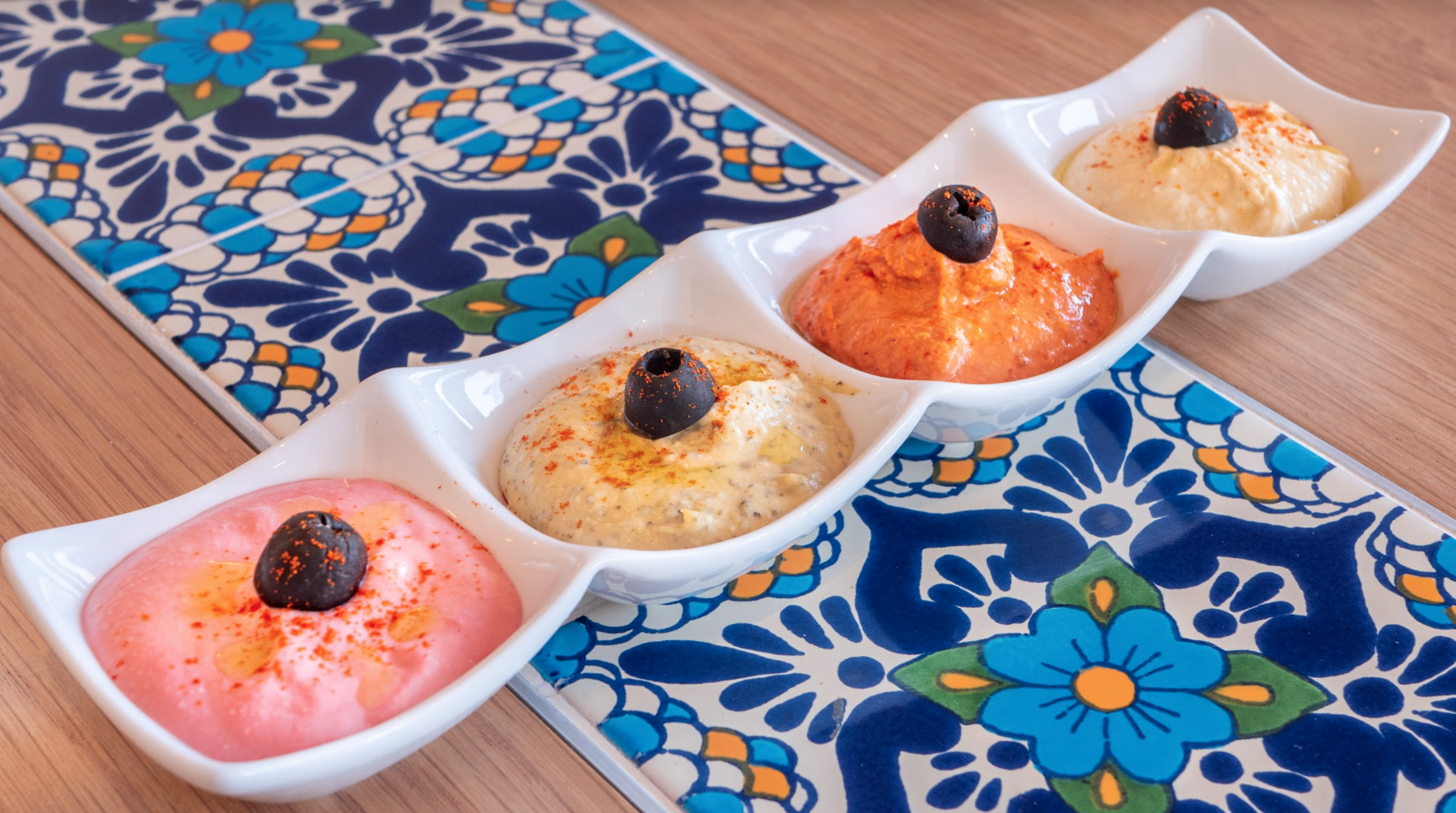 A white dish with four different types of hummus garnished with black olives, on a decorative blue and white tile table.