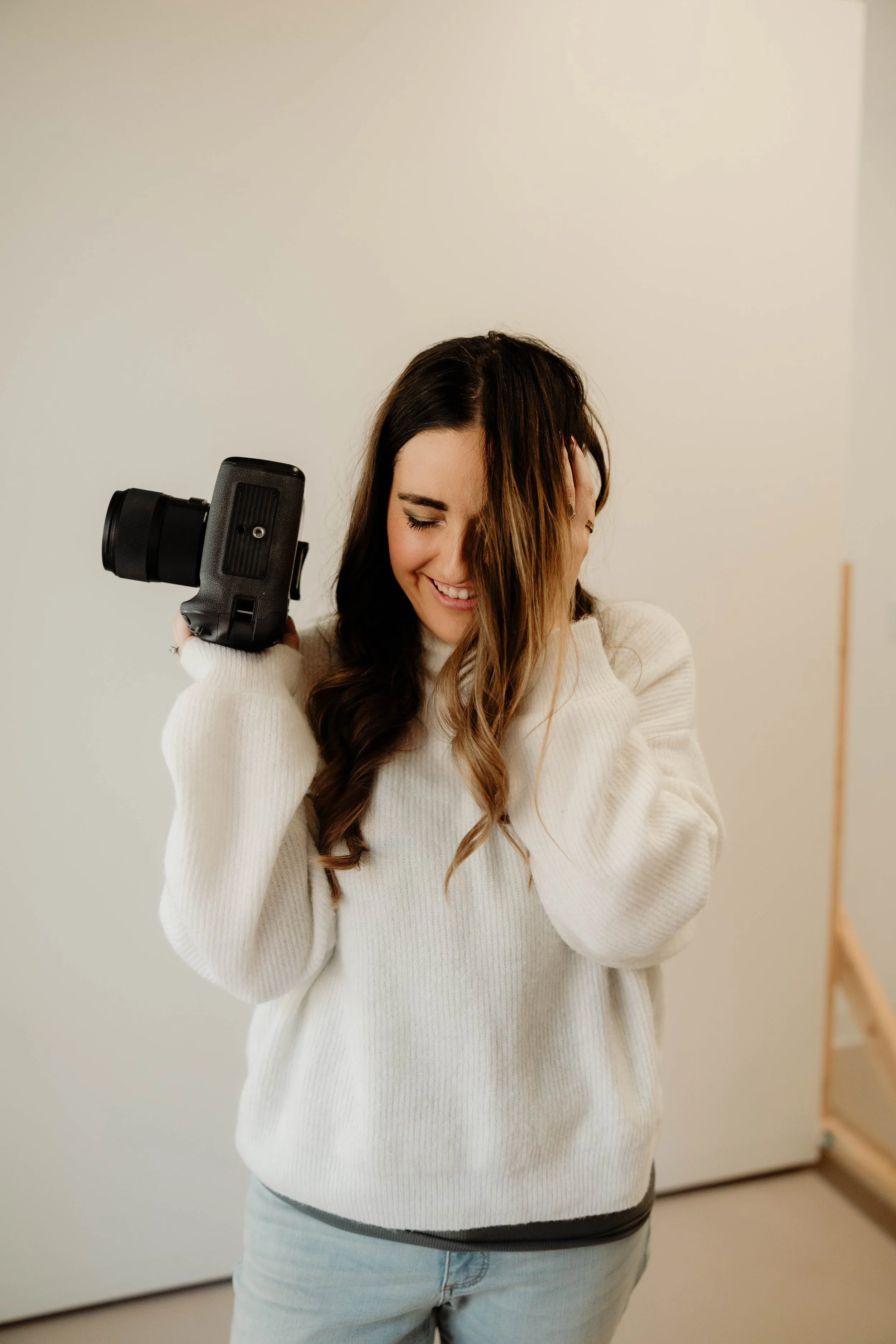 Happy woman with long brown hair holding a camera on her shoulder, wearing a white sweater, standing indoors.