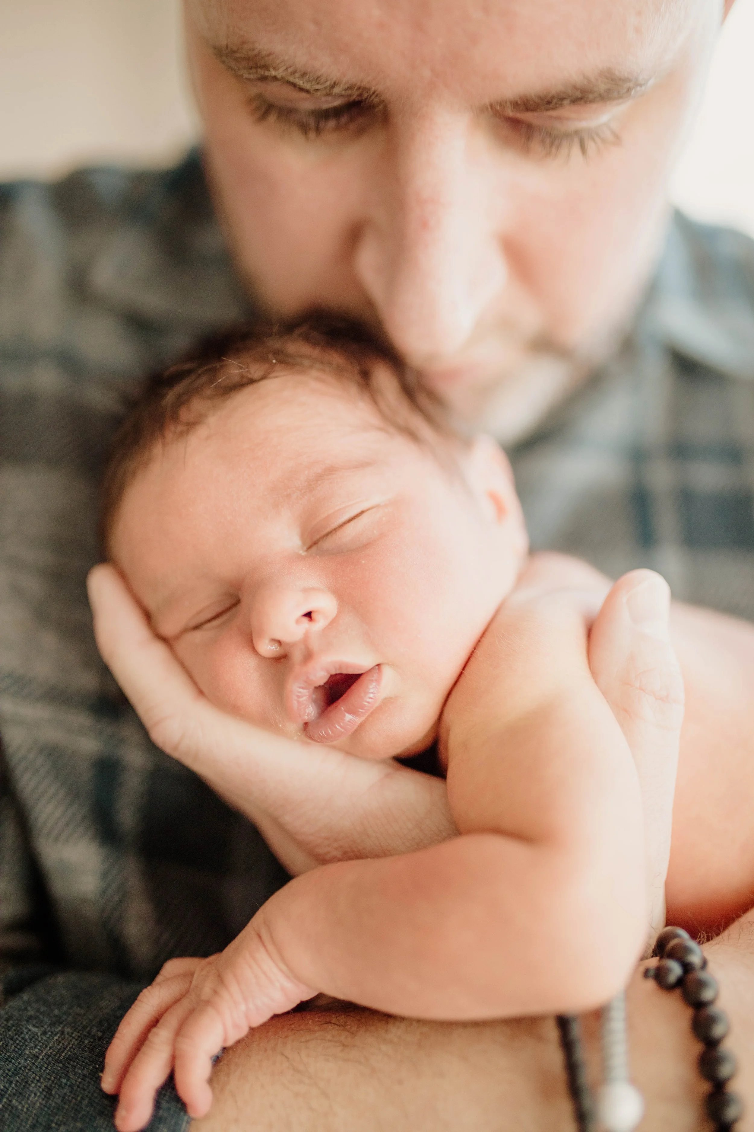 A man holding a sleeping newborn baby close to his face, cradling the baby in his hands, with the man gently kissing the baby's head.