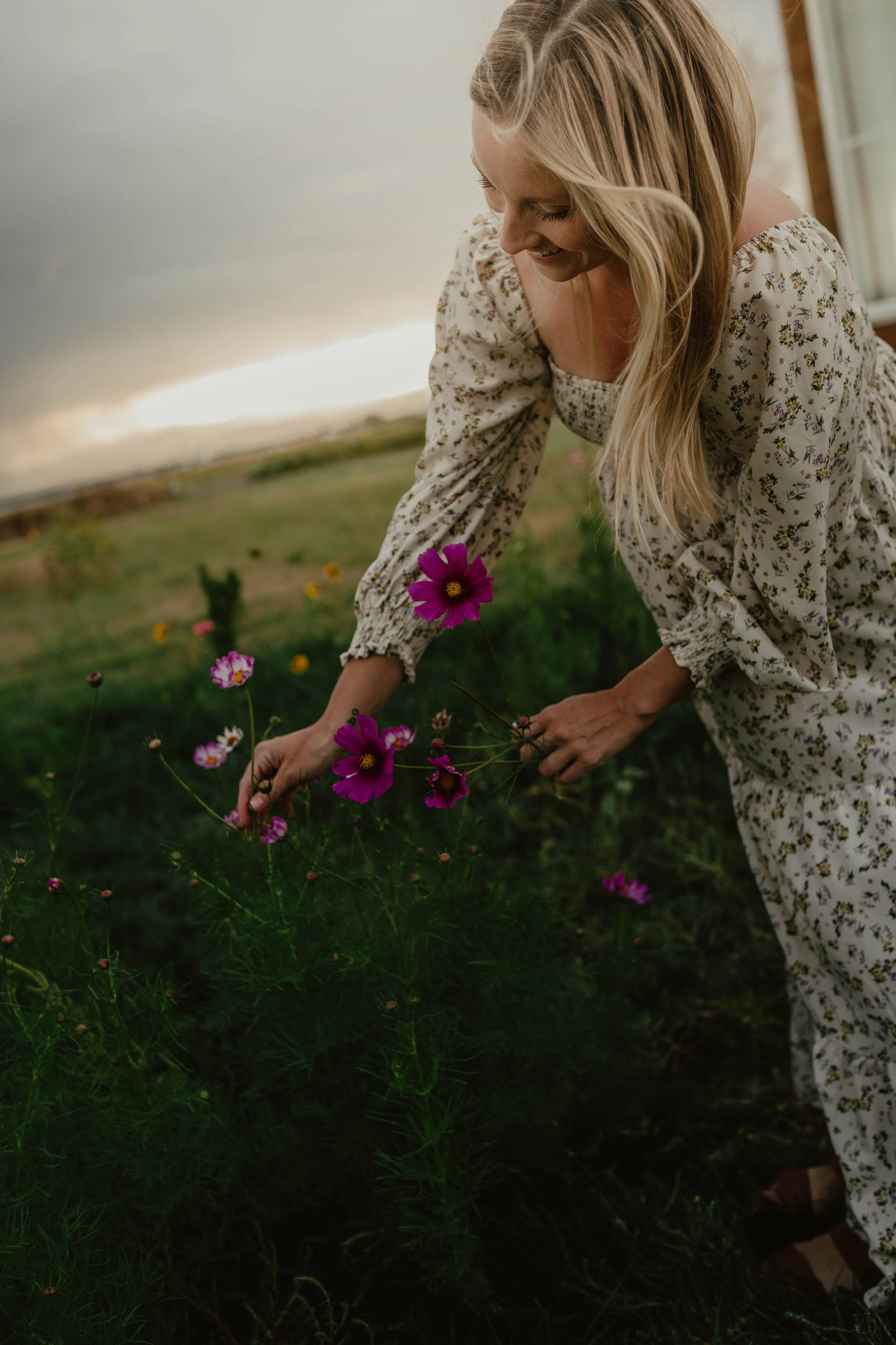 A woman in a floral dress is bending down in a garden to pick purple flowers, with a cloudy sky in the background.