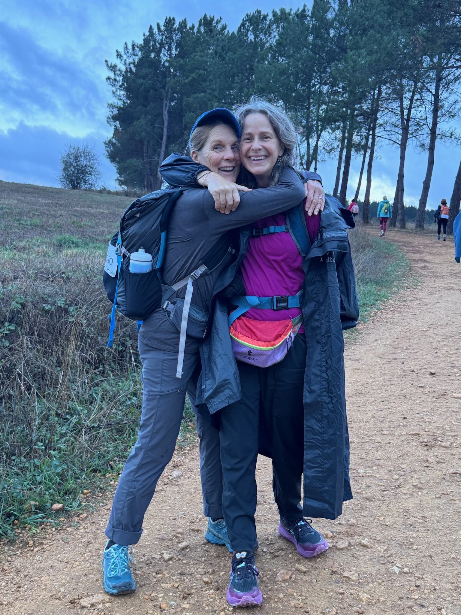 A woman is sitting on a concrete floor reading a book or notebook, while another woman is standing in front of her with her hands covering her face. They are outside a building with wooden doors and windows, shadows of a woven hanging decoration are cast on the wall, and there are palm trees in the background.