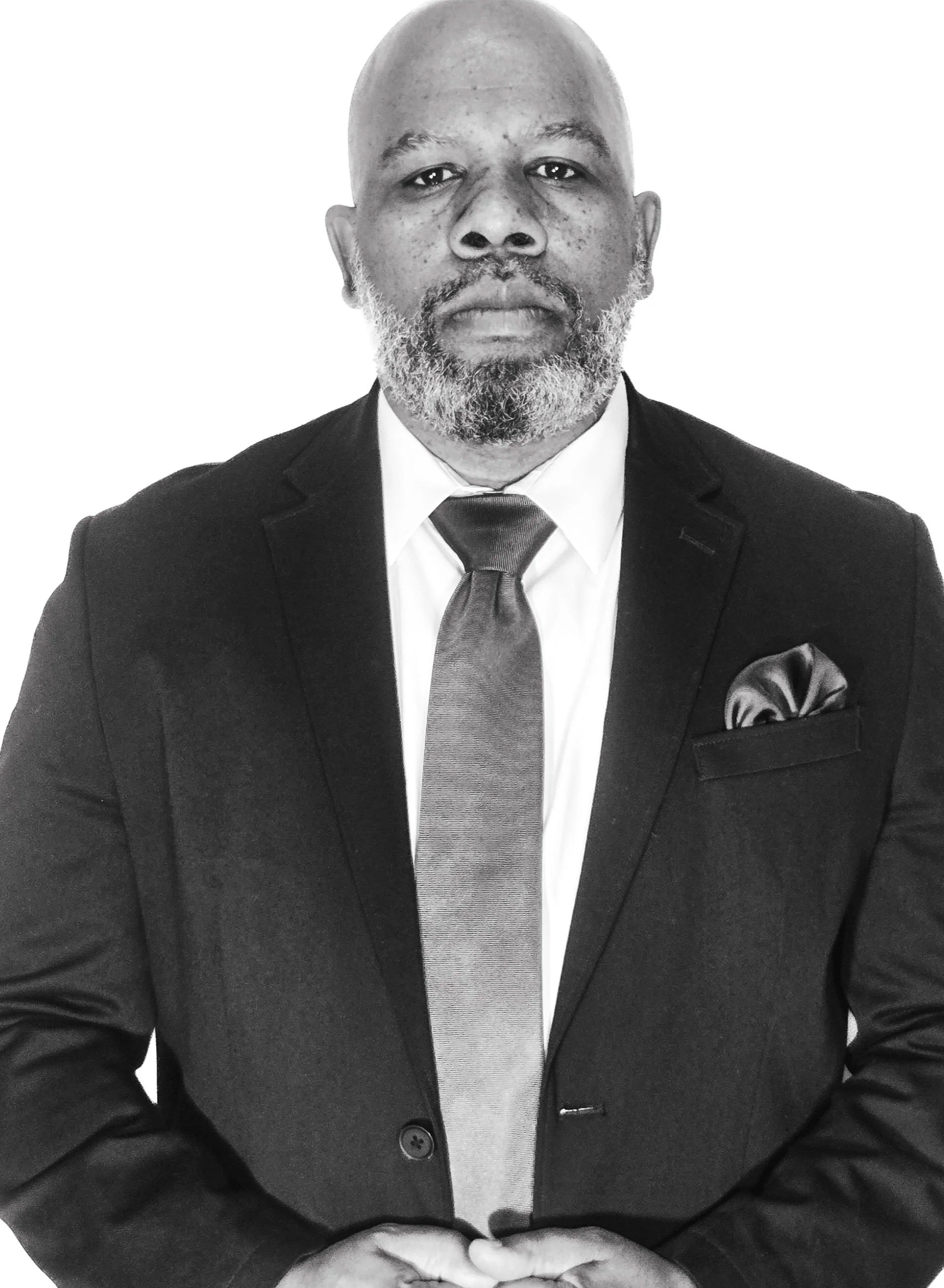 An African American man with a beard dressed in a black suit, white shirt, and gray tie, standing against a plain white background.