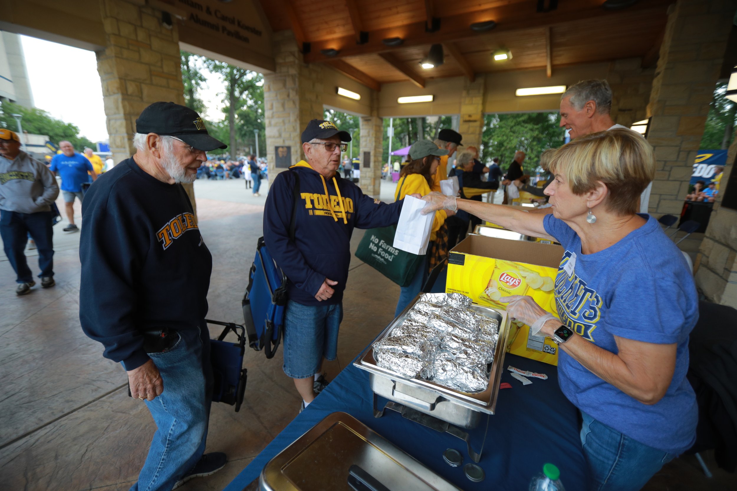 Volunteers handing out hot dogs and chips to visitors at a football tailgate in the Alumni Pavilion