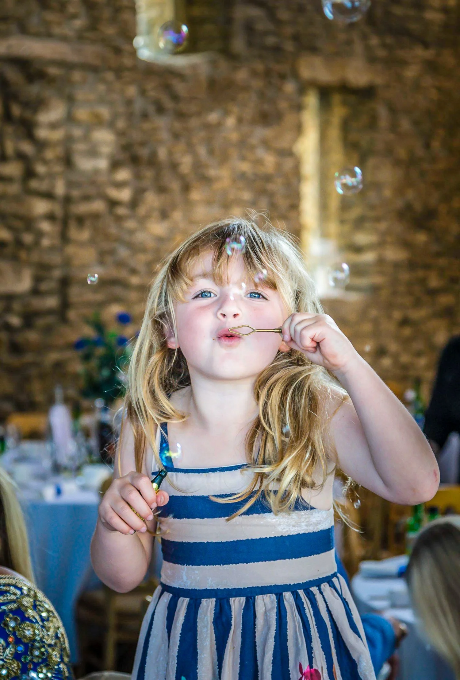 A young girl with long blonde hair and blue eyes blowing bubbles inside a room with a rustic stone wall background during a celebration or gathering.