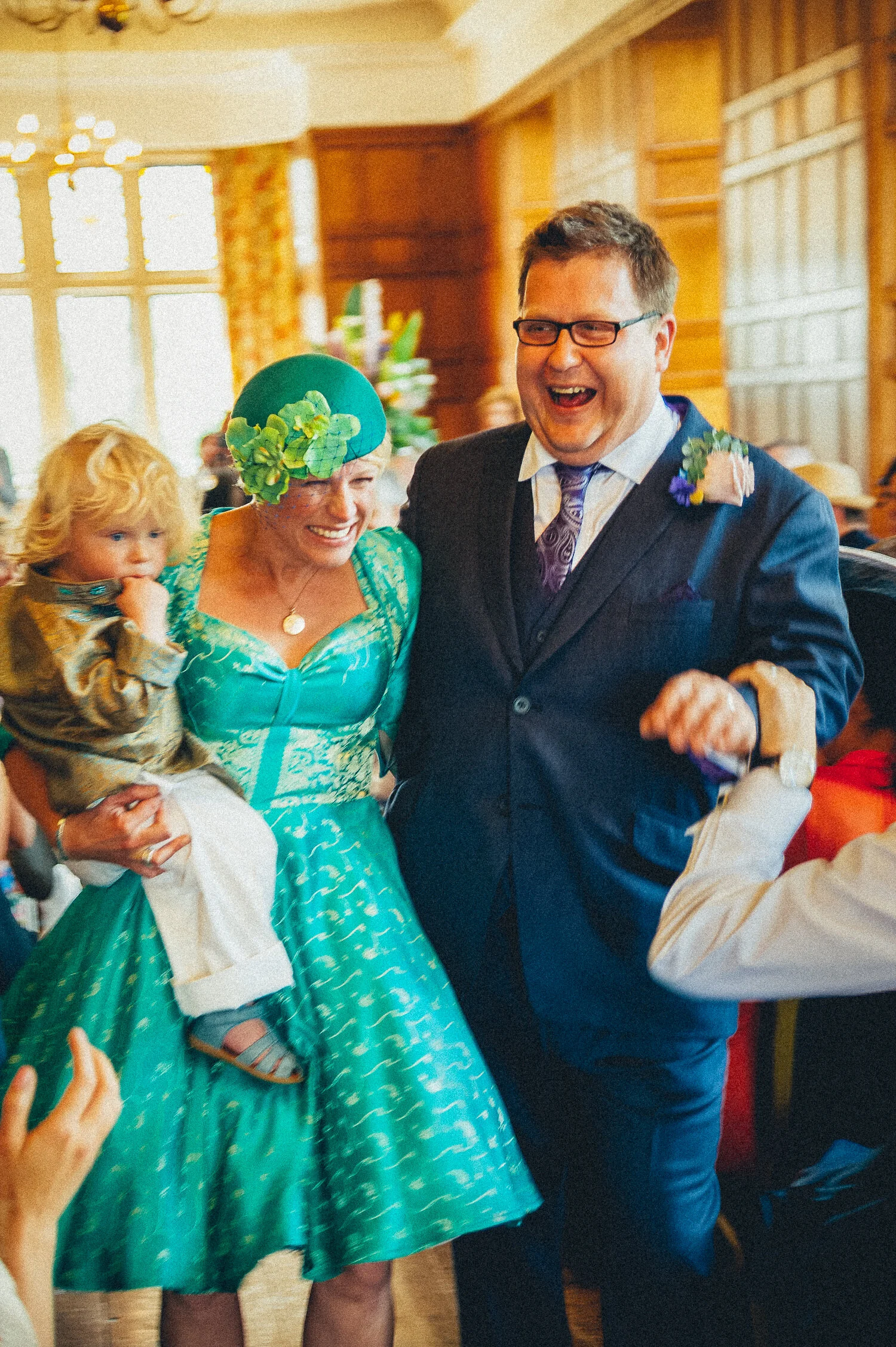People smiling and dancing at a celebration in a decorated room, with a woman holding a child and wearing a bright teal dress and hat, and a man in a suit with a boutonnière.