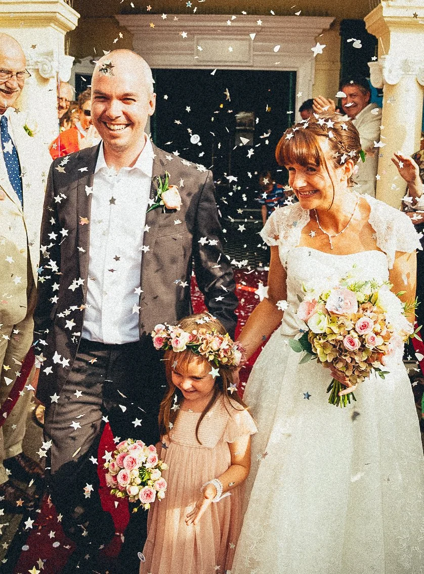 A wedding celebration with a bride, groom, and a young flower girl walking through confetti. The bride is holding a large bouquet, and the groom is smiling. Guests are visible in the background cheering.