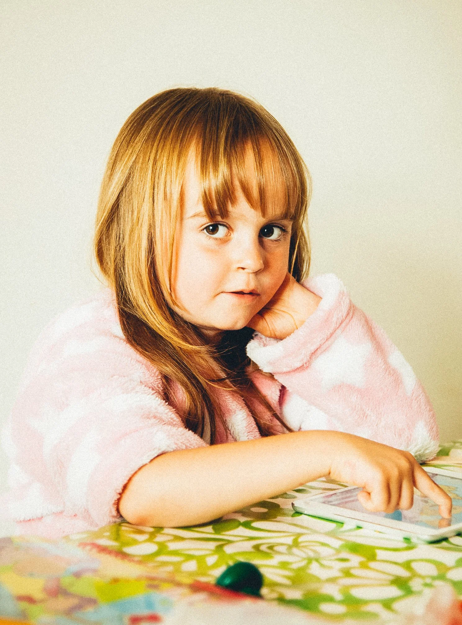 A young girl with red hair and big eyes, wearing a pink and white fuzzy sweater, sitting at a table with colorful patterned paper and engaging with her tablet.