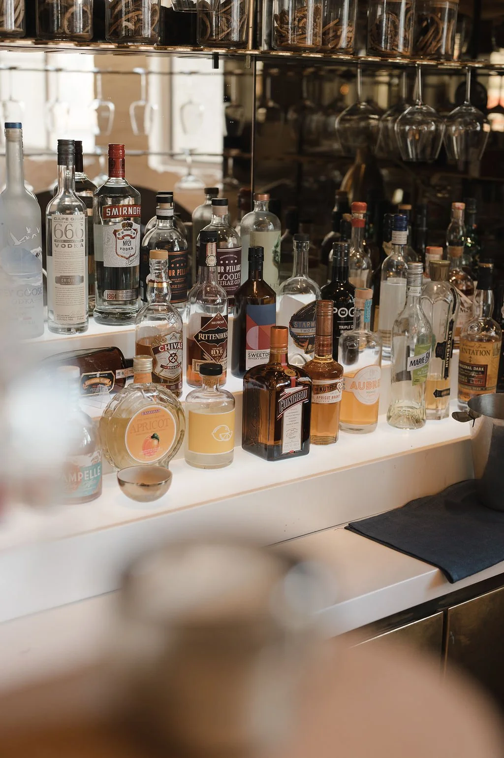A bar counter with various bottles of alcohol, including vodka, whiskey, and liqueurs, displayed on a white surface with glasses and a mirror reflecting the bottles.