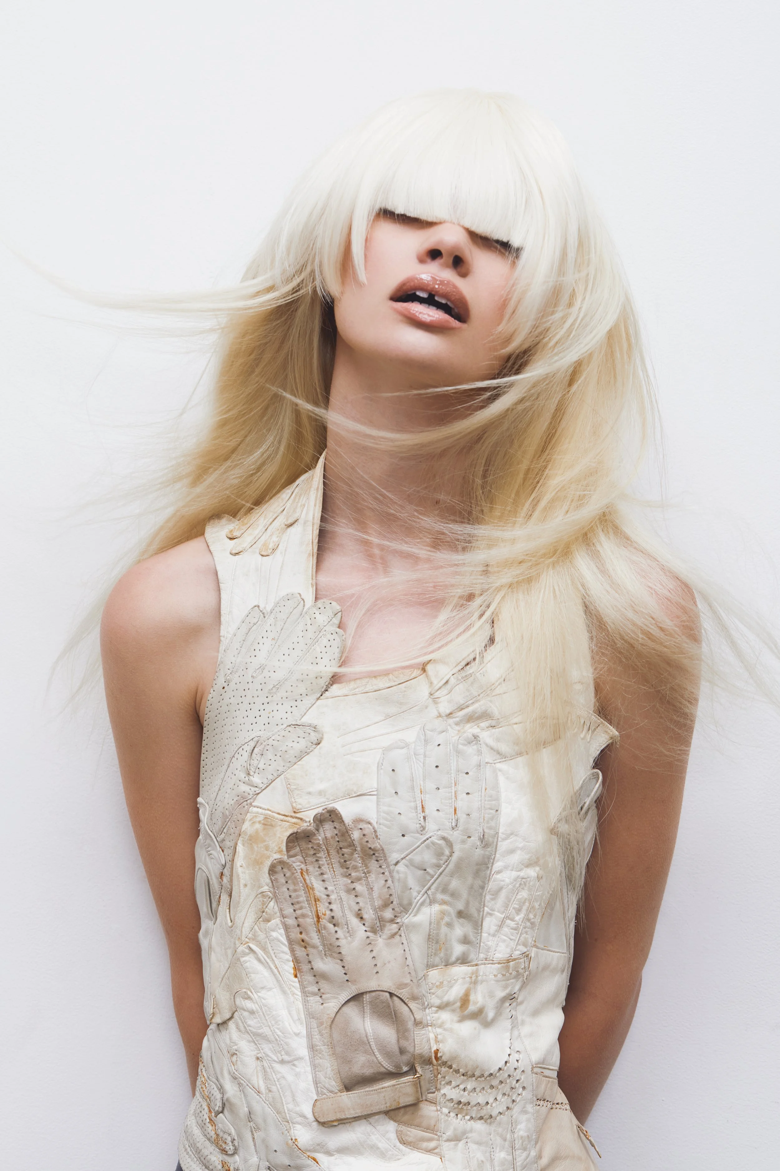 A woman with platinum blonde hair covering her eyes, wearing a sleeveless beige top, and holding her hand against her chest against a white background.