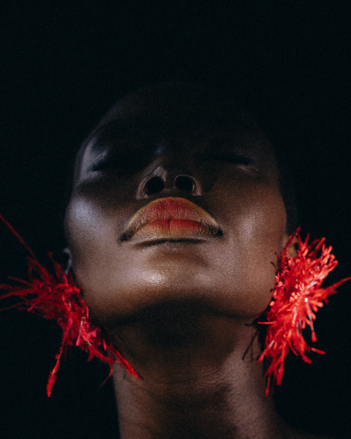 A close-up portrait of a woman with dark skin, wearing bold red feather earrings and gold lipstick, looking upwards with a dark background.