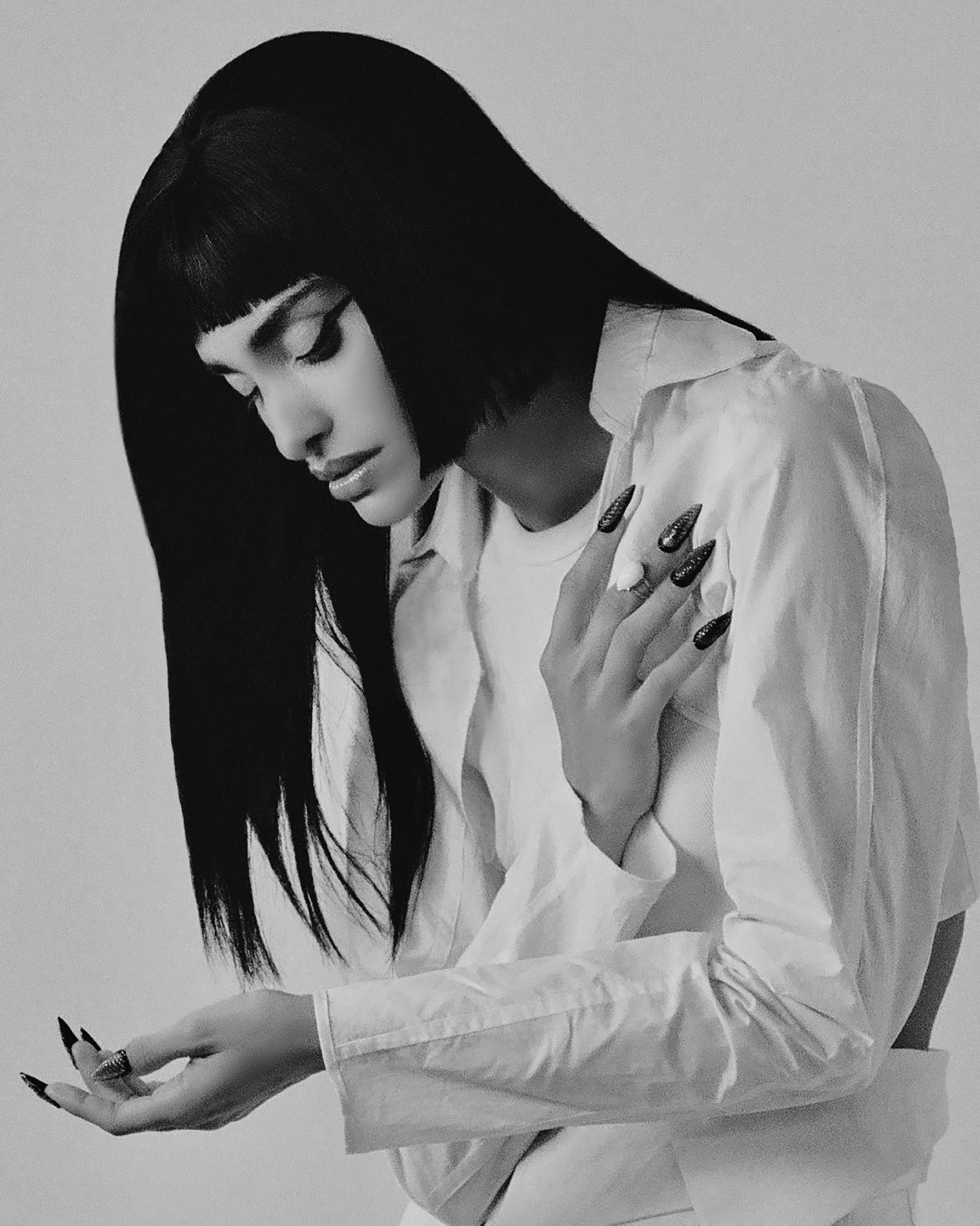 Black and white photo of a woman with long dark hair and bangs, wearing a white shirt, looking down while holding a cellphone in her left hand and touching her shoulder with her right hand.