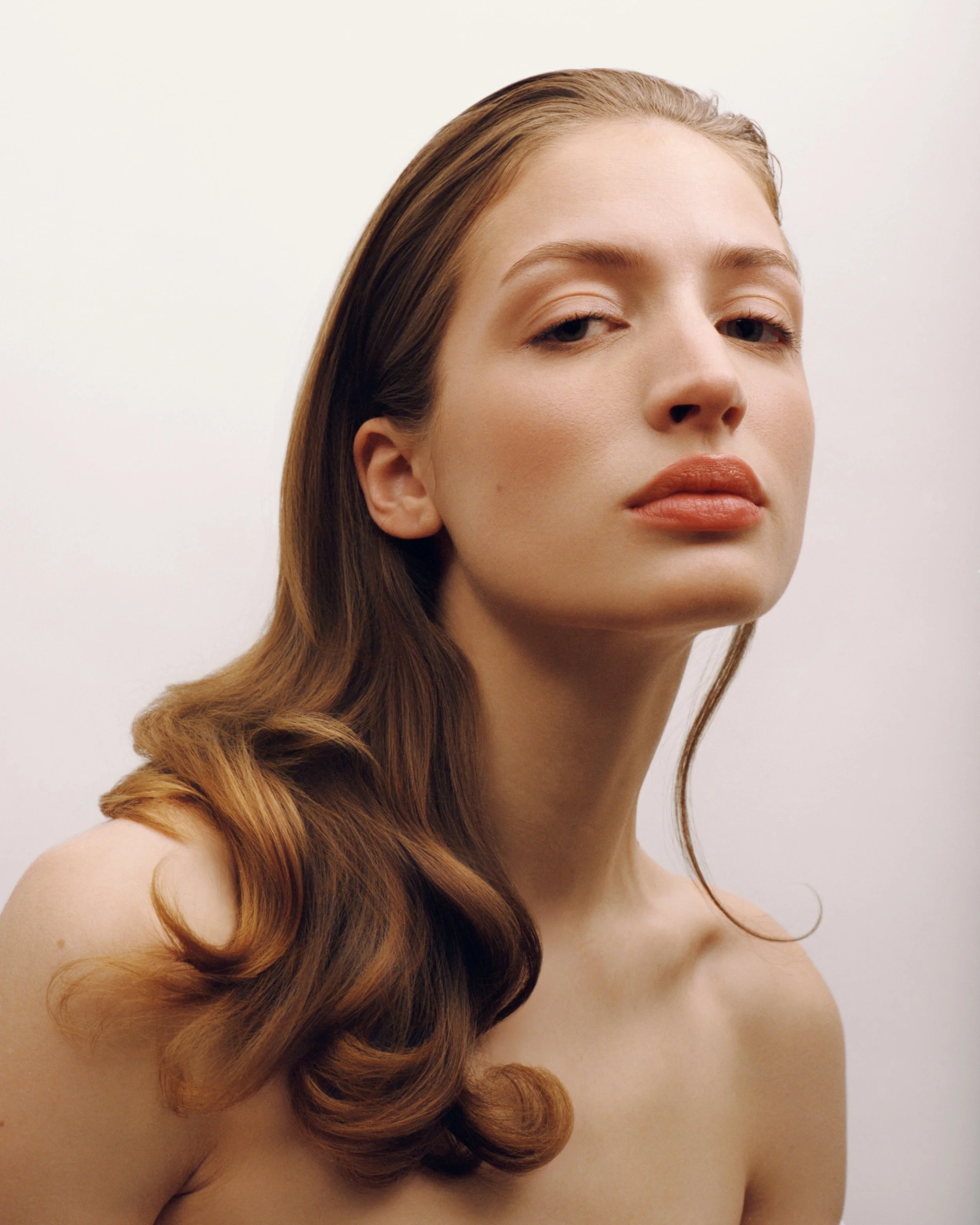 A woman with long, wavy brown hair and natural makeup, posing with a neutral expression against a plain white background.