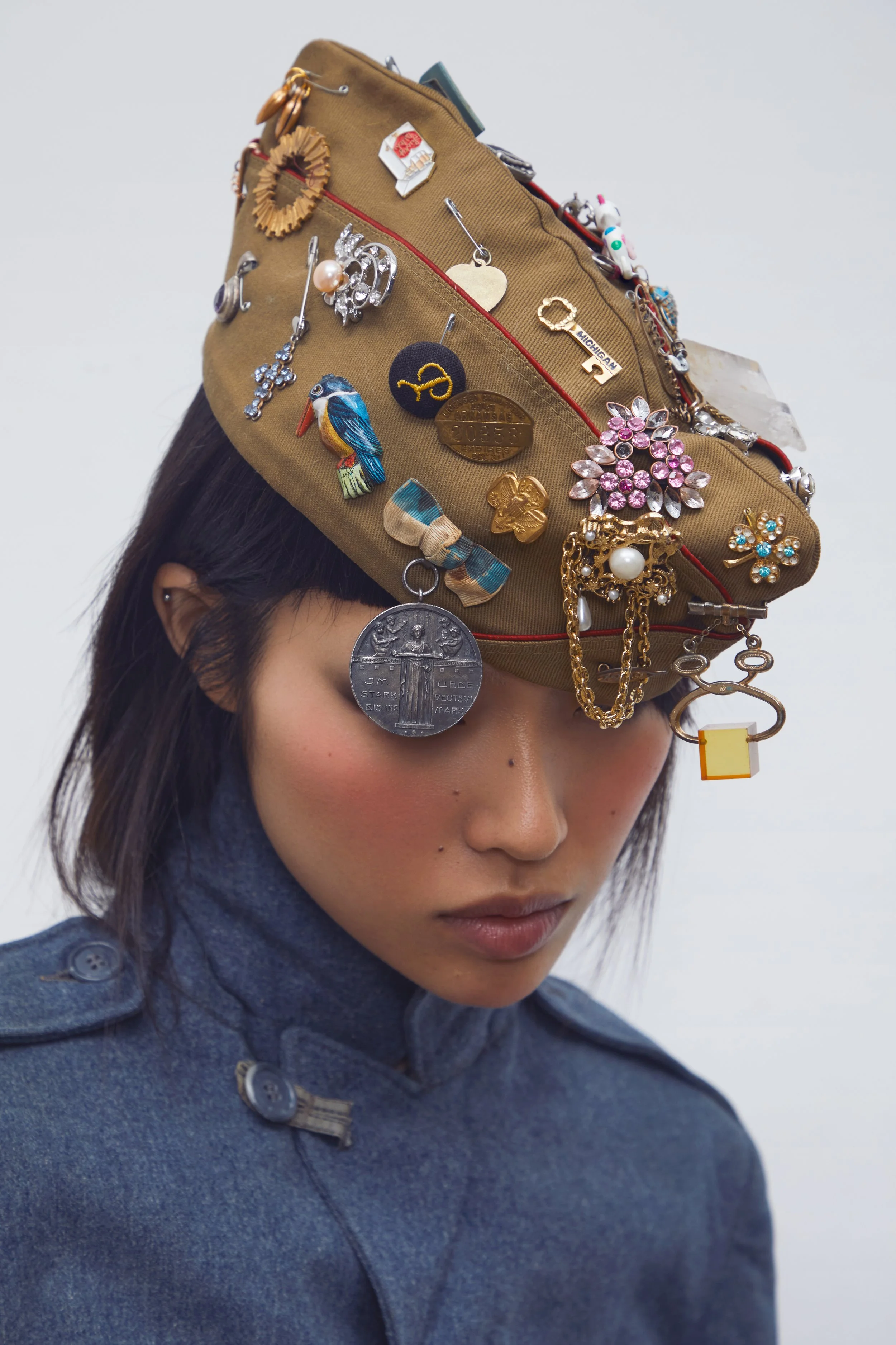 A woman wears a tan hat decorated with numerous pins, badges, and jewelry, partially covering her short dark hair. She is dressed in a blue, military-style coat with button details, her eyes gently closed, and her head tilted downward.