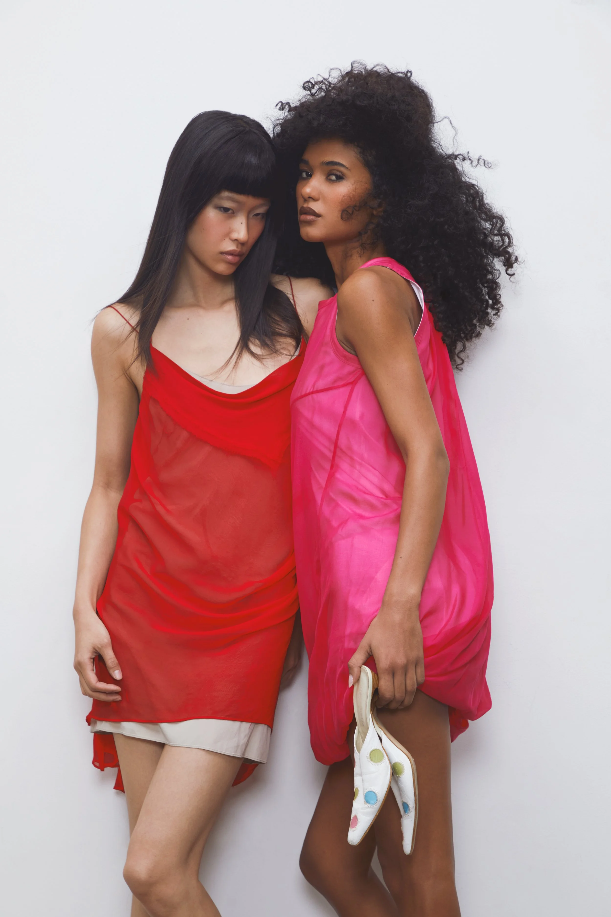 Two women standing close together against a white wall, both wearing colorful, semi-sheer dresses. The woman on the left has long straight black hair with bangs, wearing a red dress with thin straps, and appears to be looking down. The woman on the r