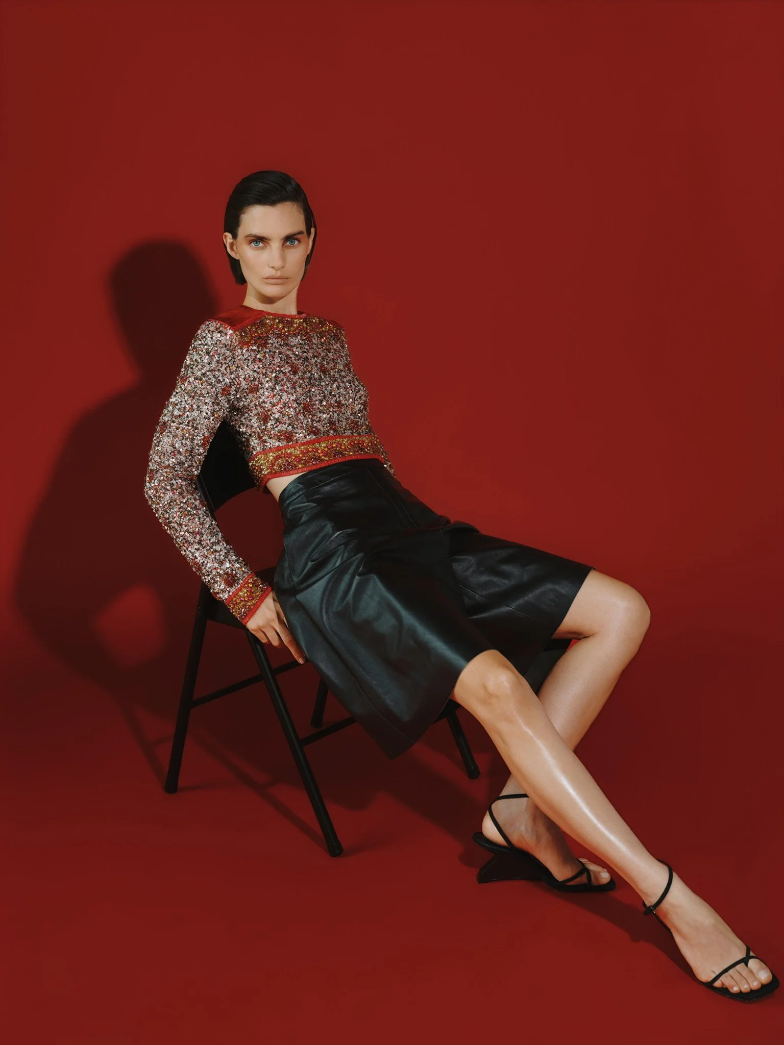 A woman with dark hair, wearing a glittery long-sleeve crop top, black leather skirt, and black strappy heels, sitting on a black chair against a red background.