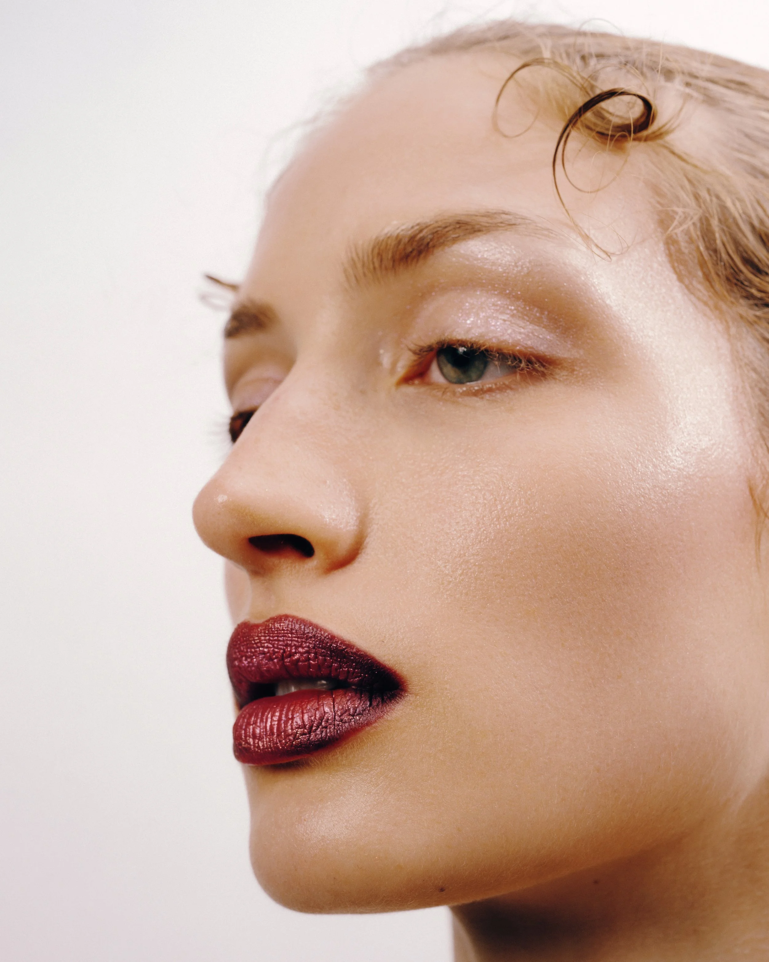Close-up of a woman with fair skin and blond hair, wearing makeup with nude eyeshadow and dark red lipstick, looking slightly to the side against a plain white background.