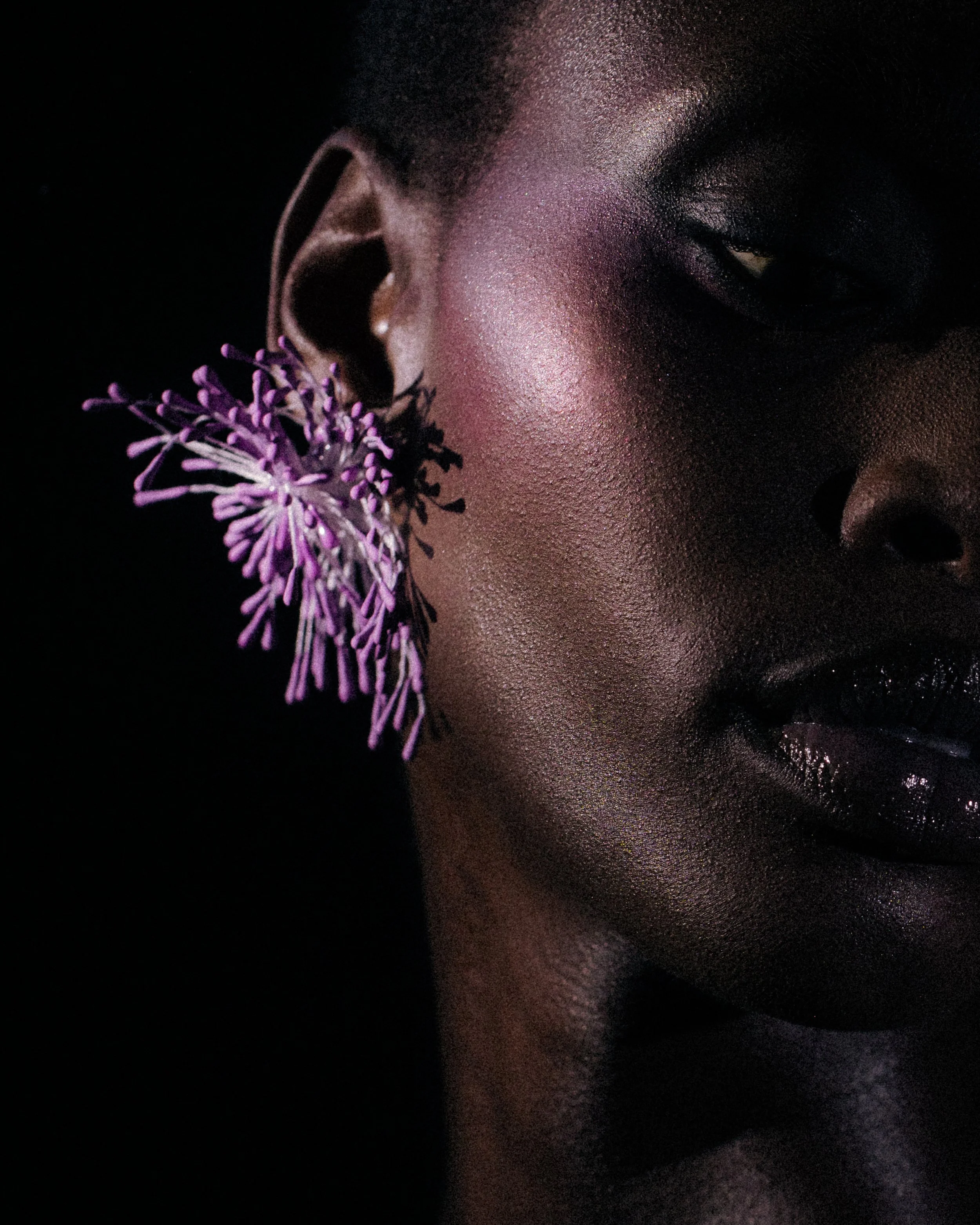 Close-up of a woman with dark skin wearing purple flower earrings, with pink eye makeup and shiny lips, against a dark background.