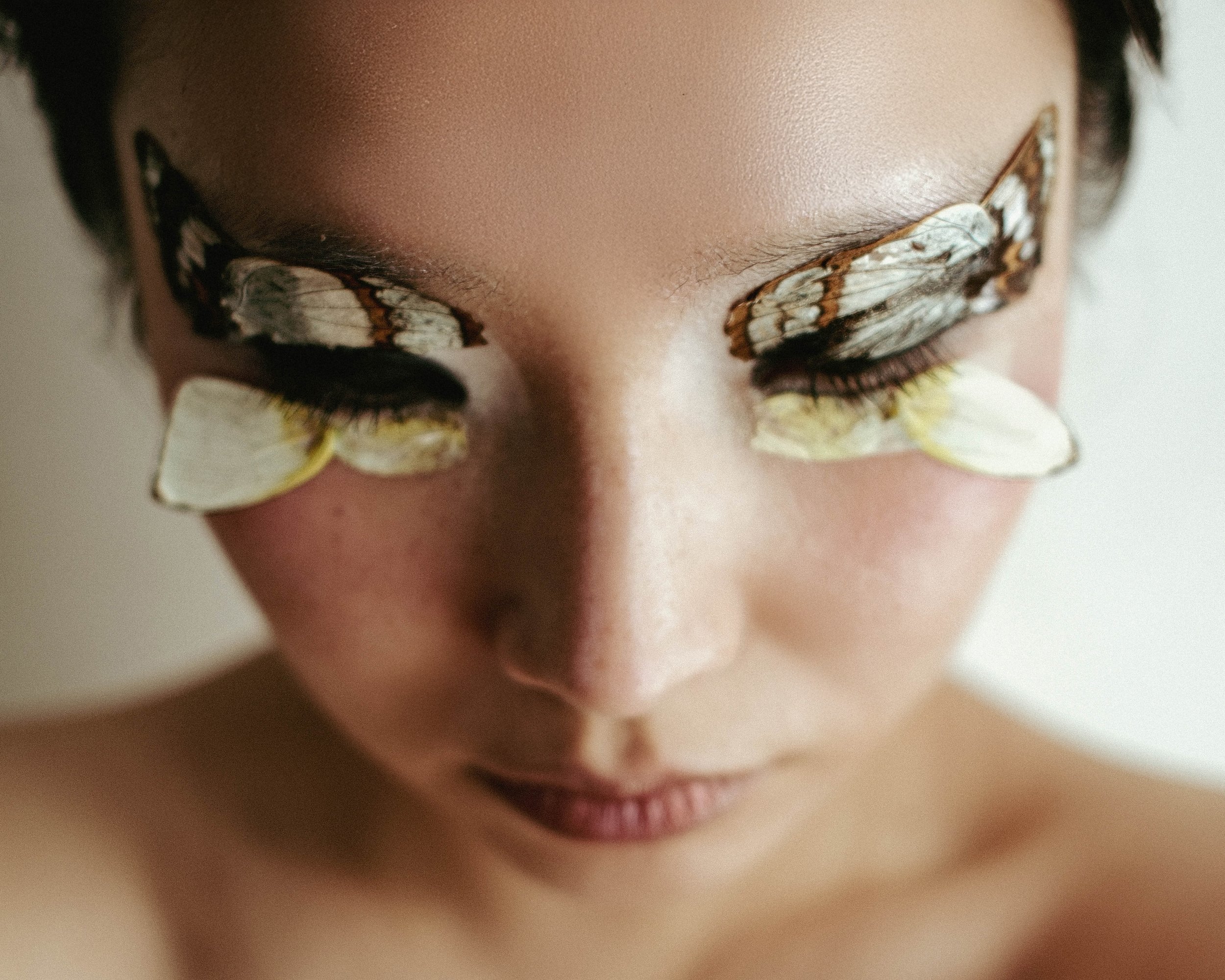 Close-up of a woman's face with butterflies on her eyelids and cheeks, her eyes closed, and her head slightly tilted downward.