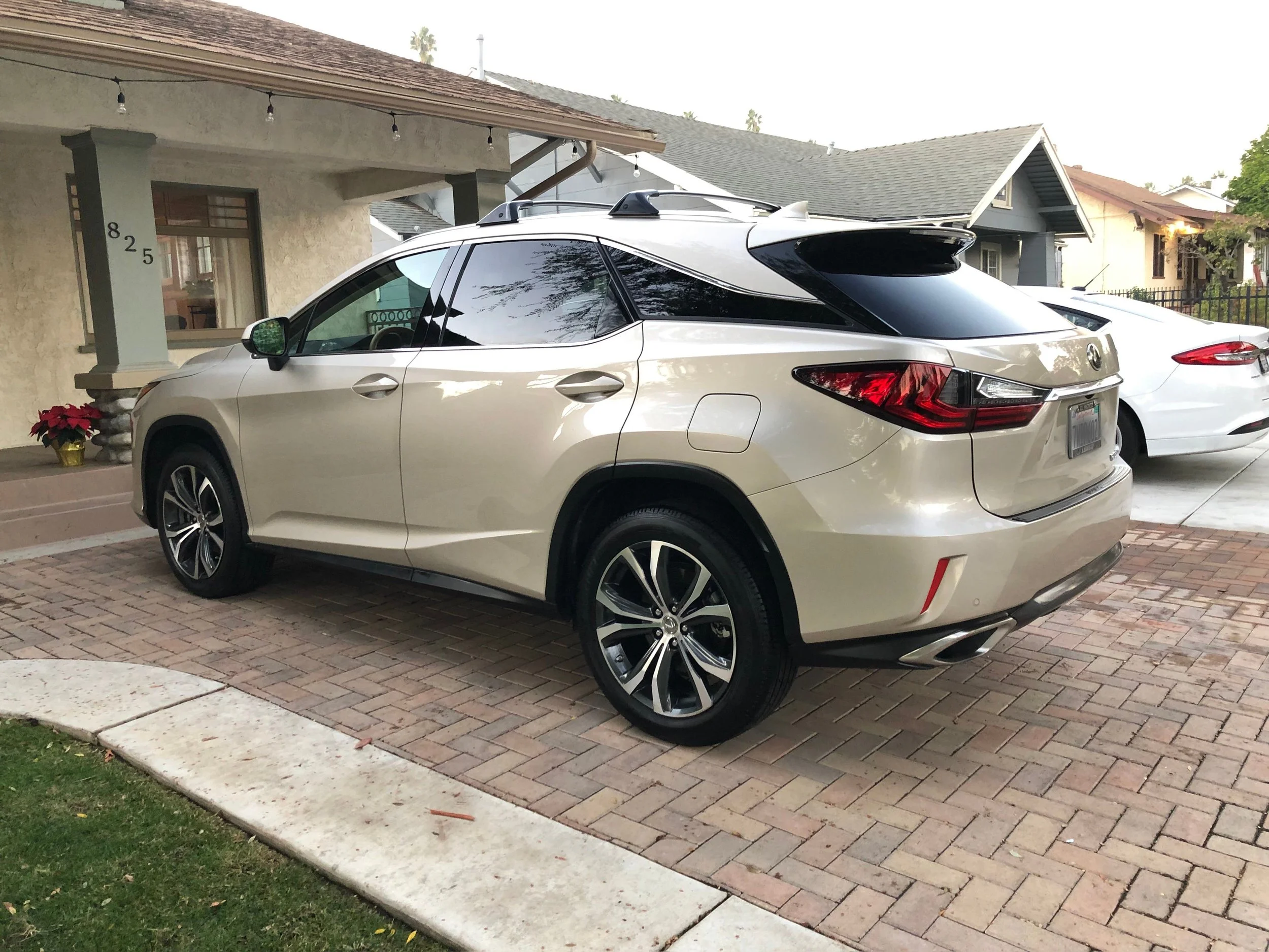 Silver SUV parked in front of a house on a brick driveway, with another white car parked nearby and houses visible in the background.