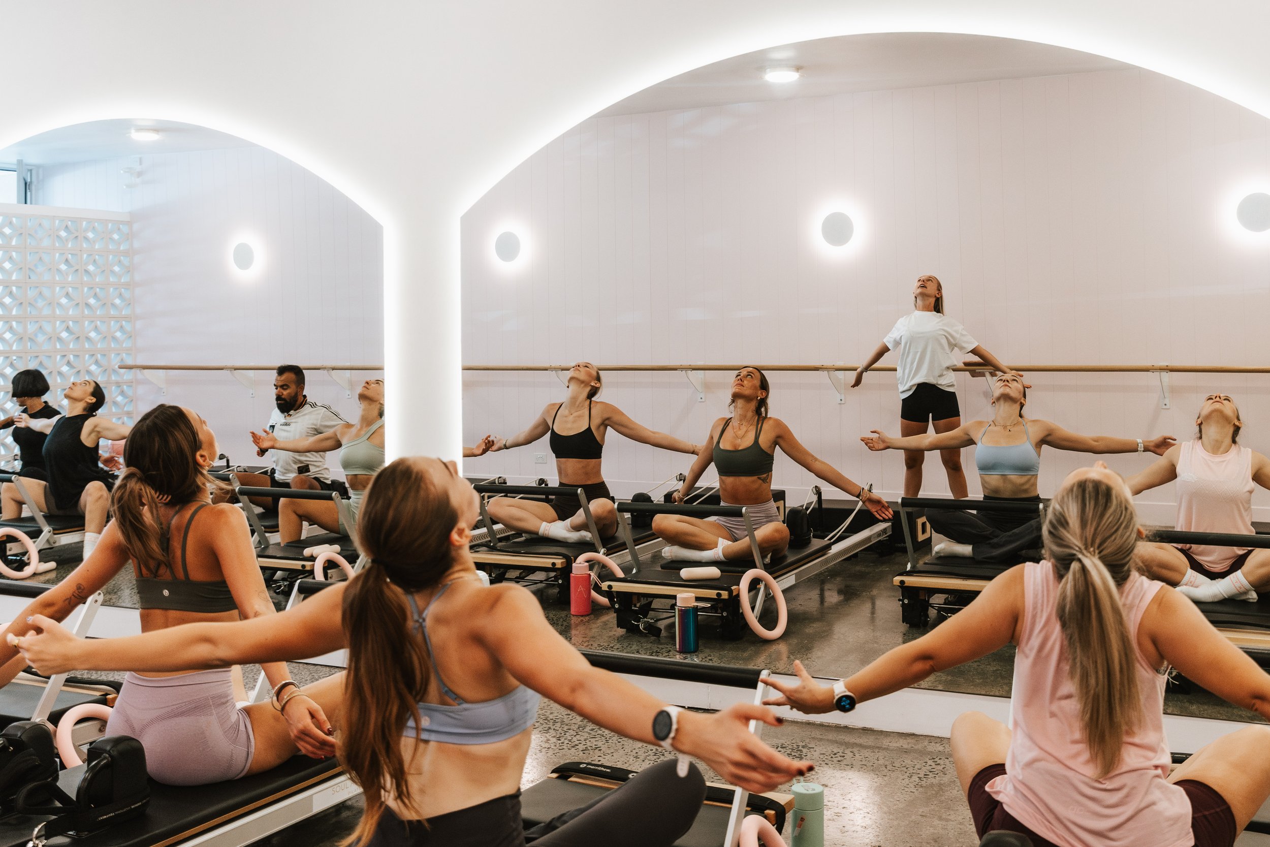 People participating in a fitness class or yoga session in a spacious, well-lit room with white walls and a barre, performing stretching or meditation exercises, some seated and others standing.