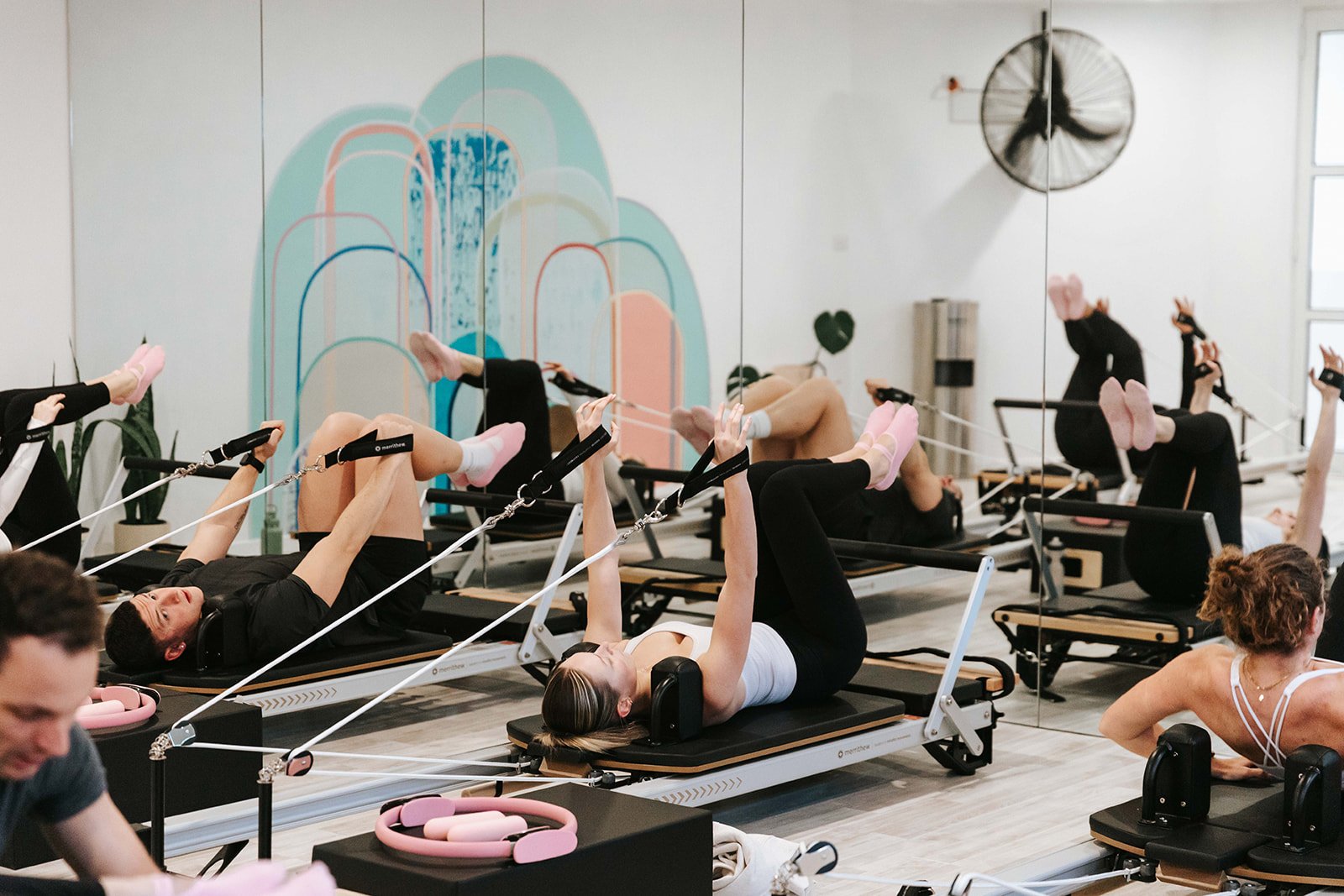 People participating in a Pilates class using reformer machines in a studio with white walls, a colorful wall mural, and a mirrored wall.