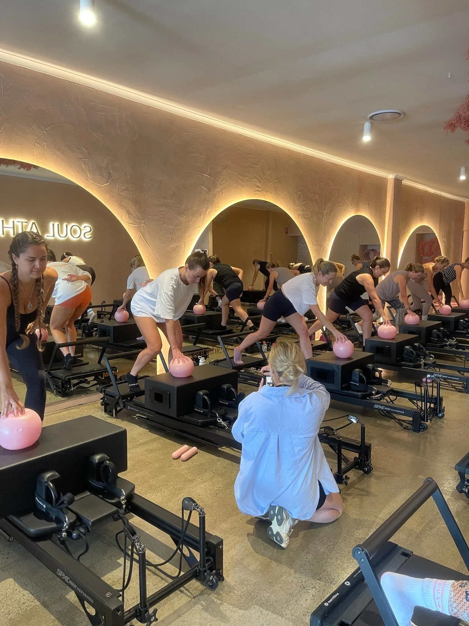 Women participating in a reformer Pilates class in a fitness studio, using pink kettlebells on reformer machines, with an instructor kneeling in front taking photos.