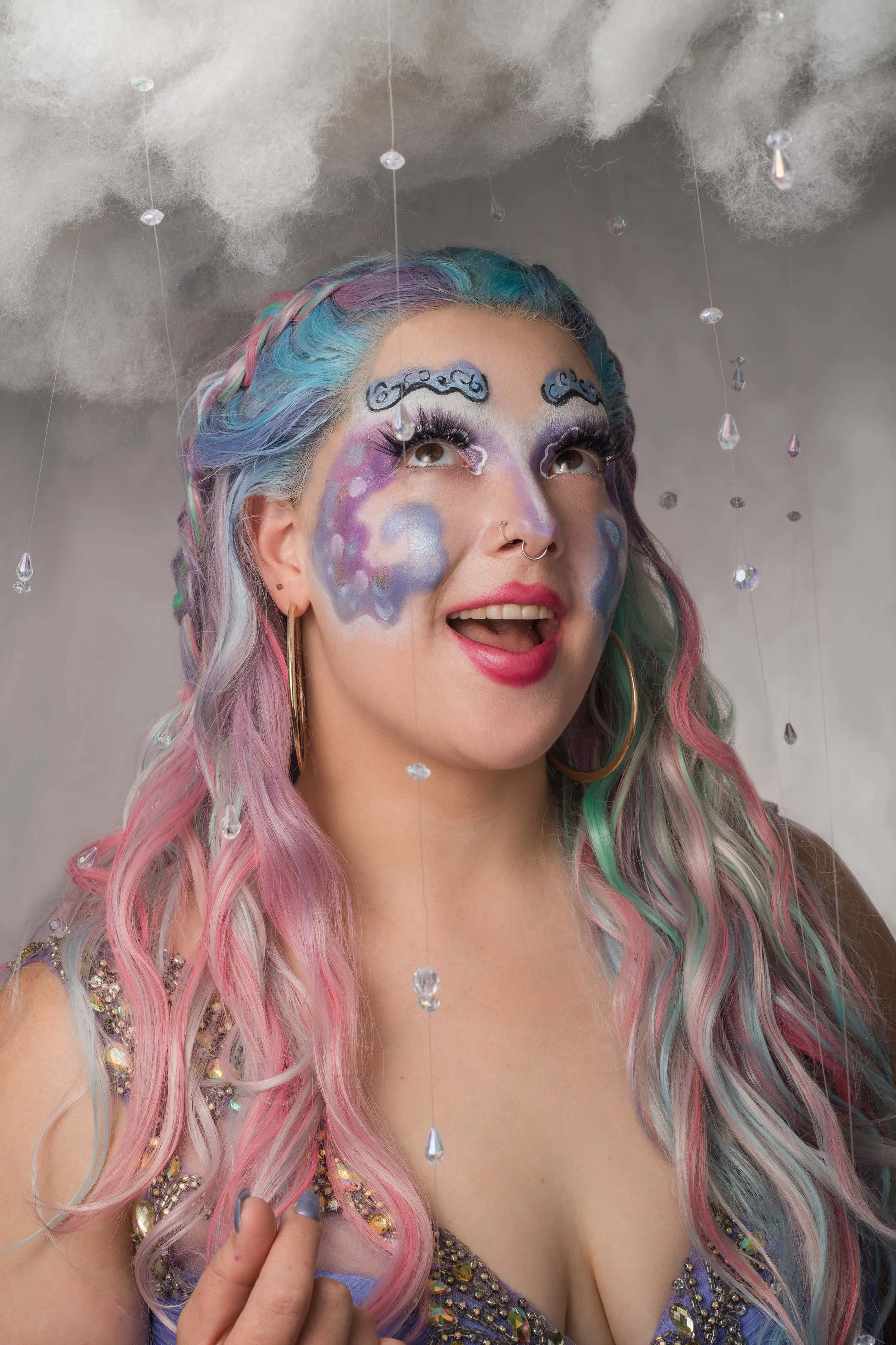 A woman with pastel rainbow hair and elaborate face makeup, dressed in a beaded top, surrounded by hanging crystal droplets and fake clouds above her head.