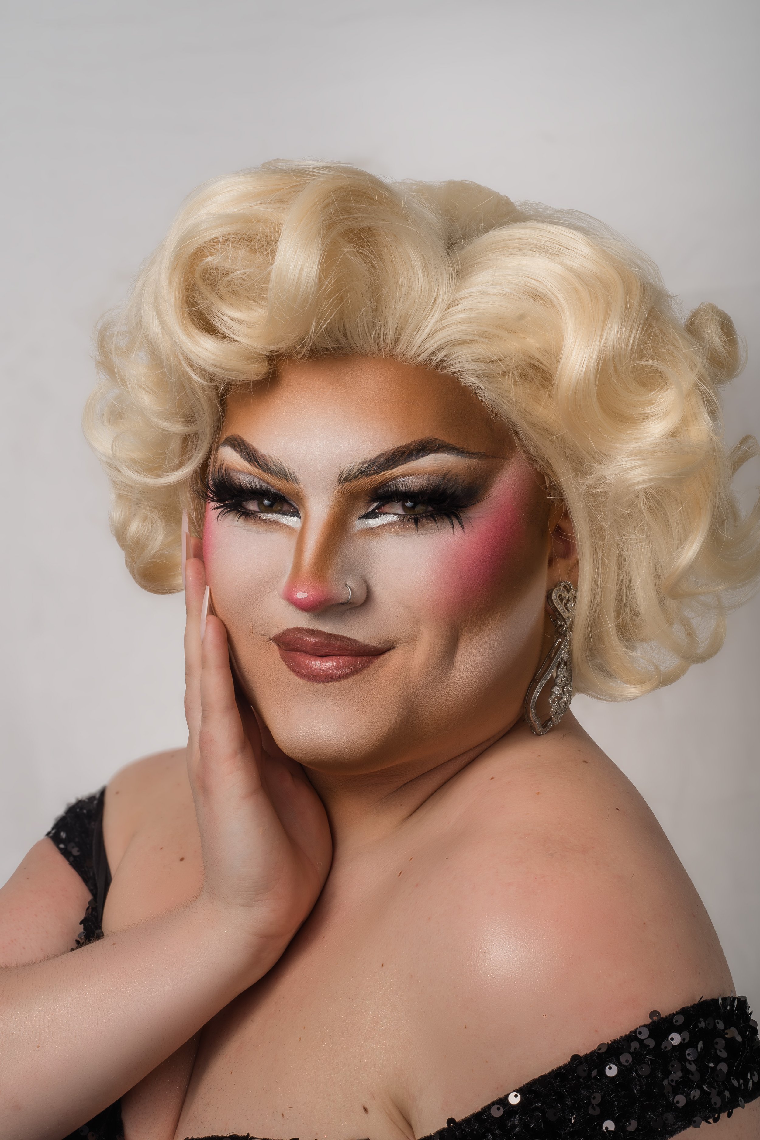 A person with platinum blonde, curly hair and heavy makeup, including dark eyeshadow, pink blush, and lip color, touching their face with a hand, dressed in a black, sequined top and wearing large, ornate earrings.