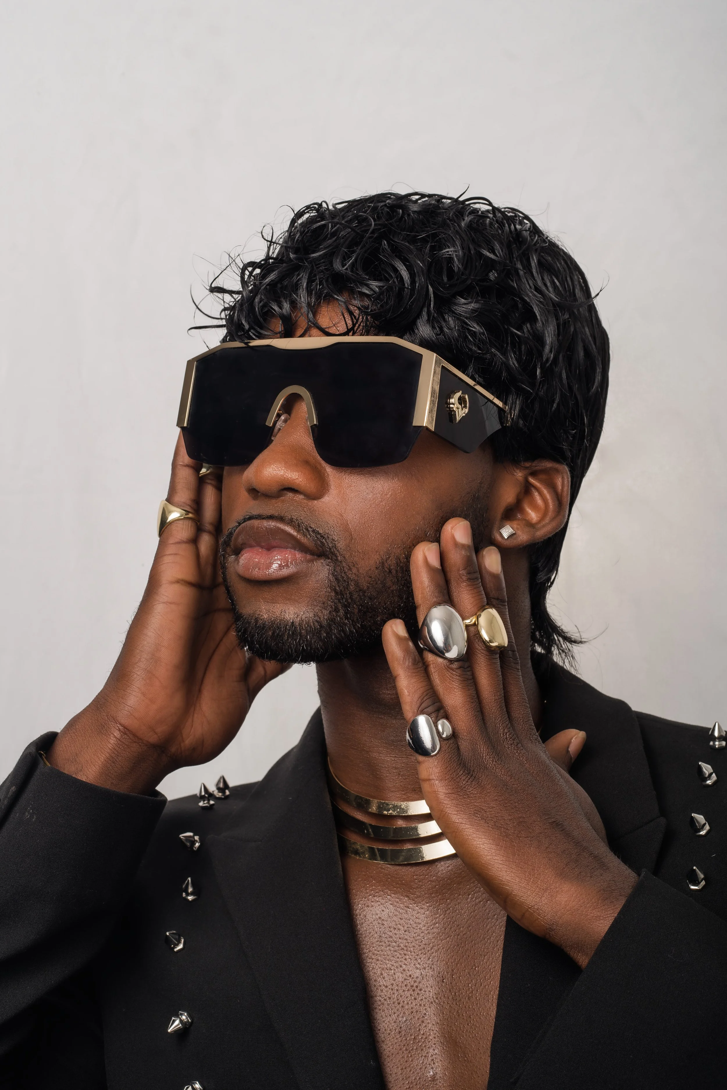 A person with dark skin, wearing oversized black sunglasses with gold accents, multiple rings on fingers, and a layered gold choker, posing against a plain background.