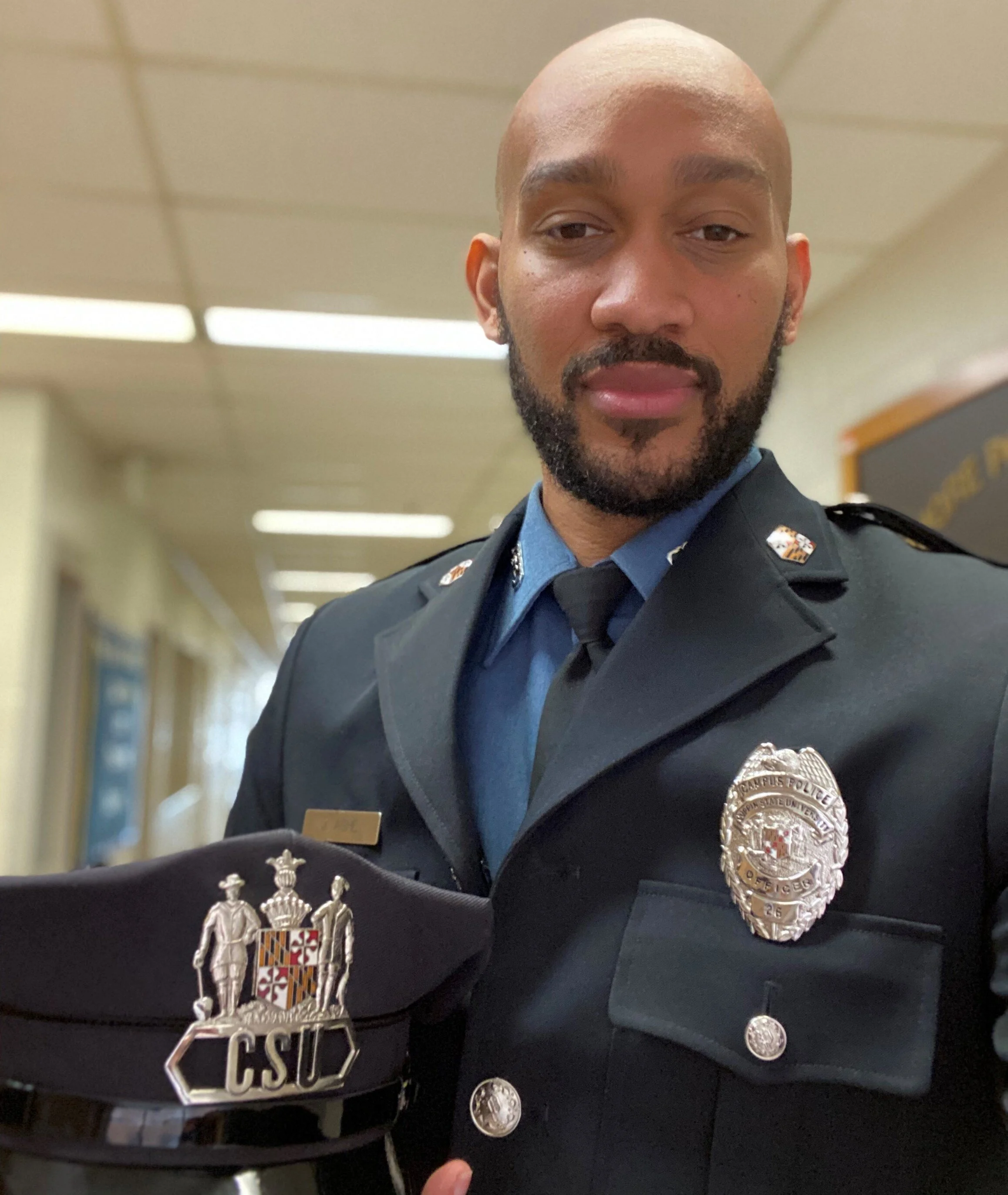 A male police officer in uniform inside a building, holding a Maryland State Police hat with a badge marked 'CSU' and a badge on his uniform's chest, posing for a photo.