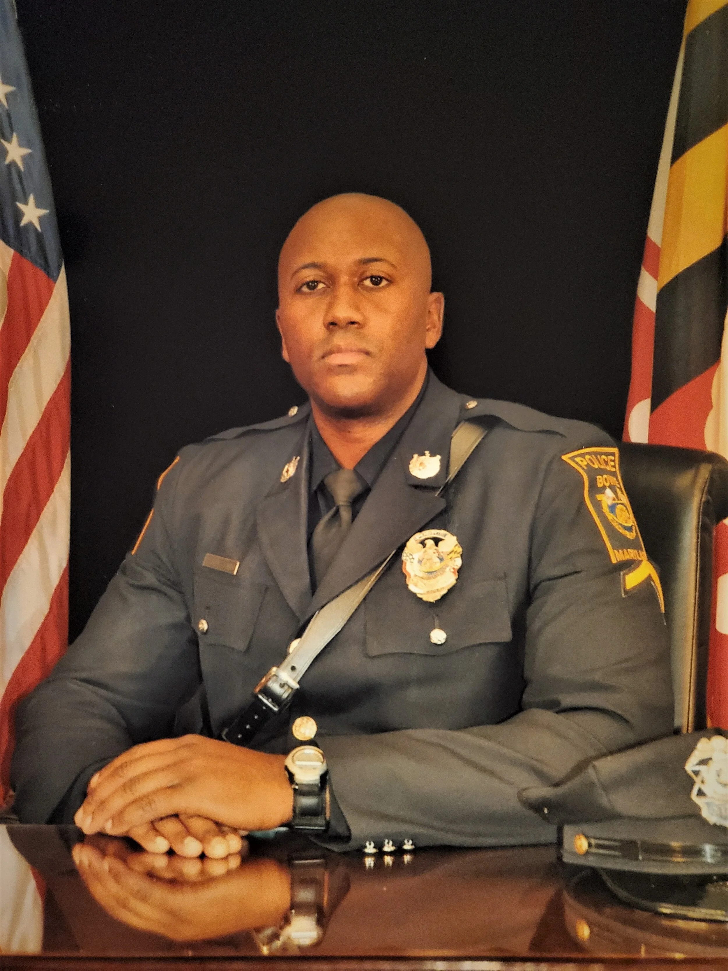 A police officer sitting at a desk with American and Maryland flags behind him, wearing a dark police uniform with badges and insignia.