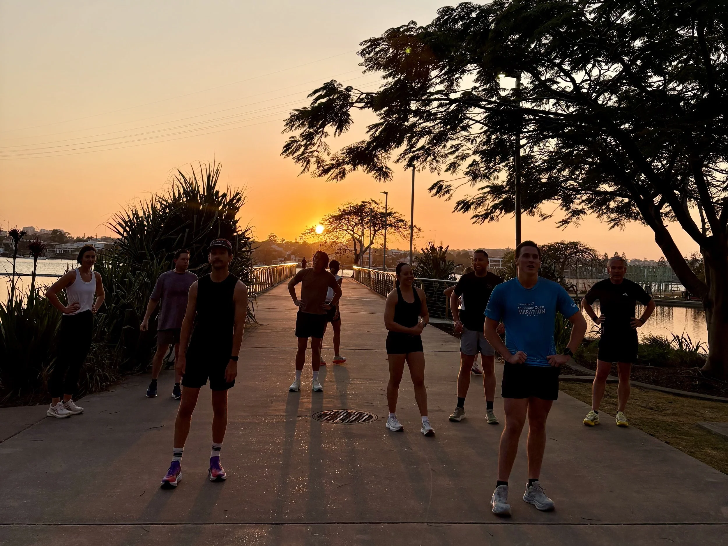 A group of people gathered outdoors during sunset, some standing and some walking, near trees and a paved walkway.