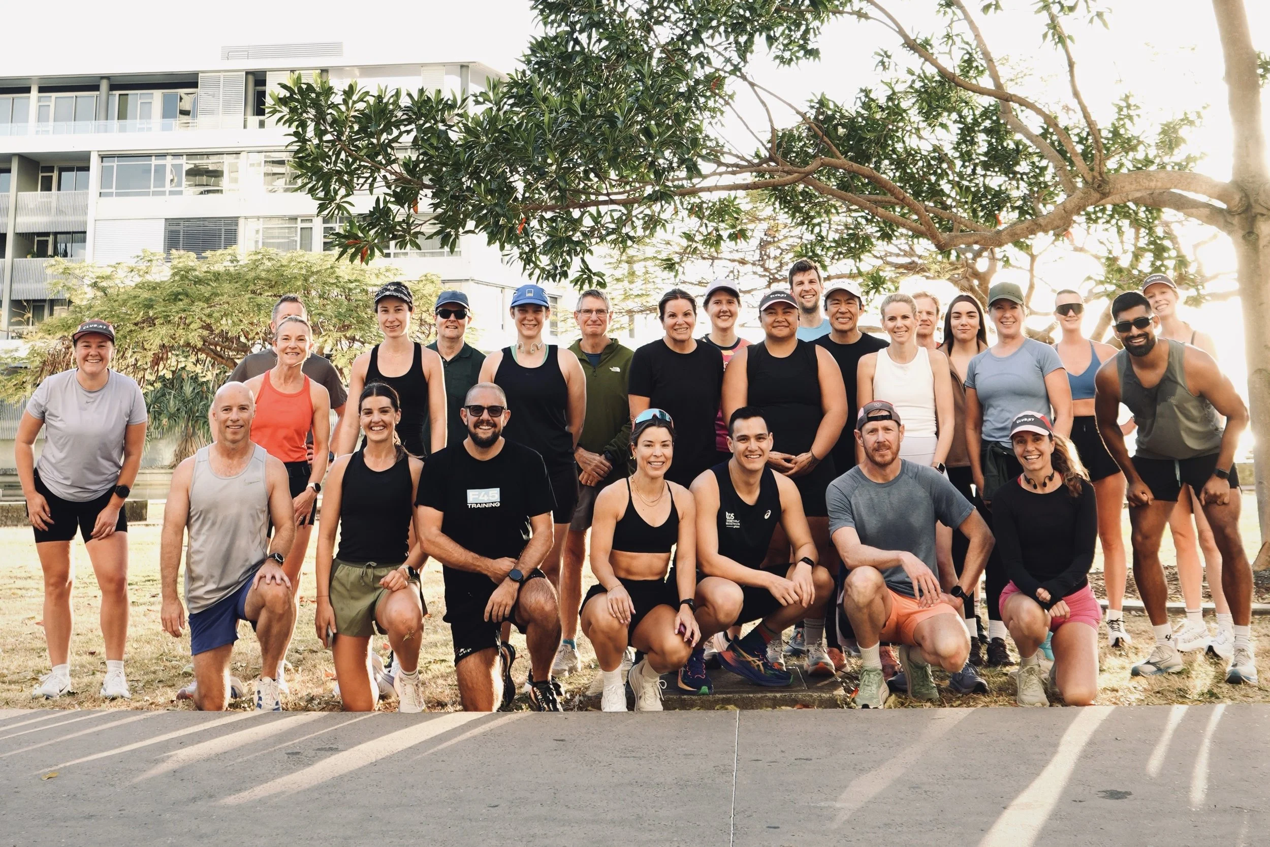 A large group of people gathered outdoors during sunset, posing for a photo on a grassy area with trees and modern buildings in the background. Some individuals are wearing athletic clothing and caps, and a sign with the word 'SWEAT' is visible in front of the group.