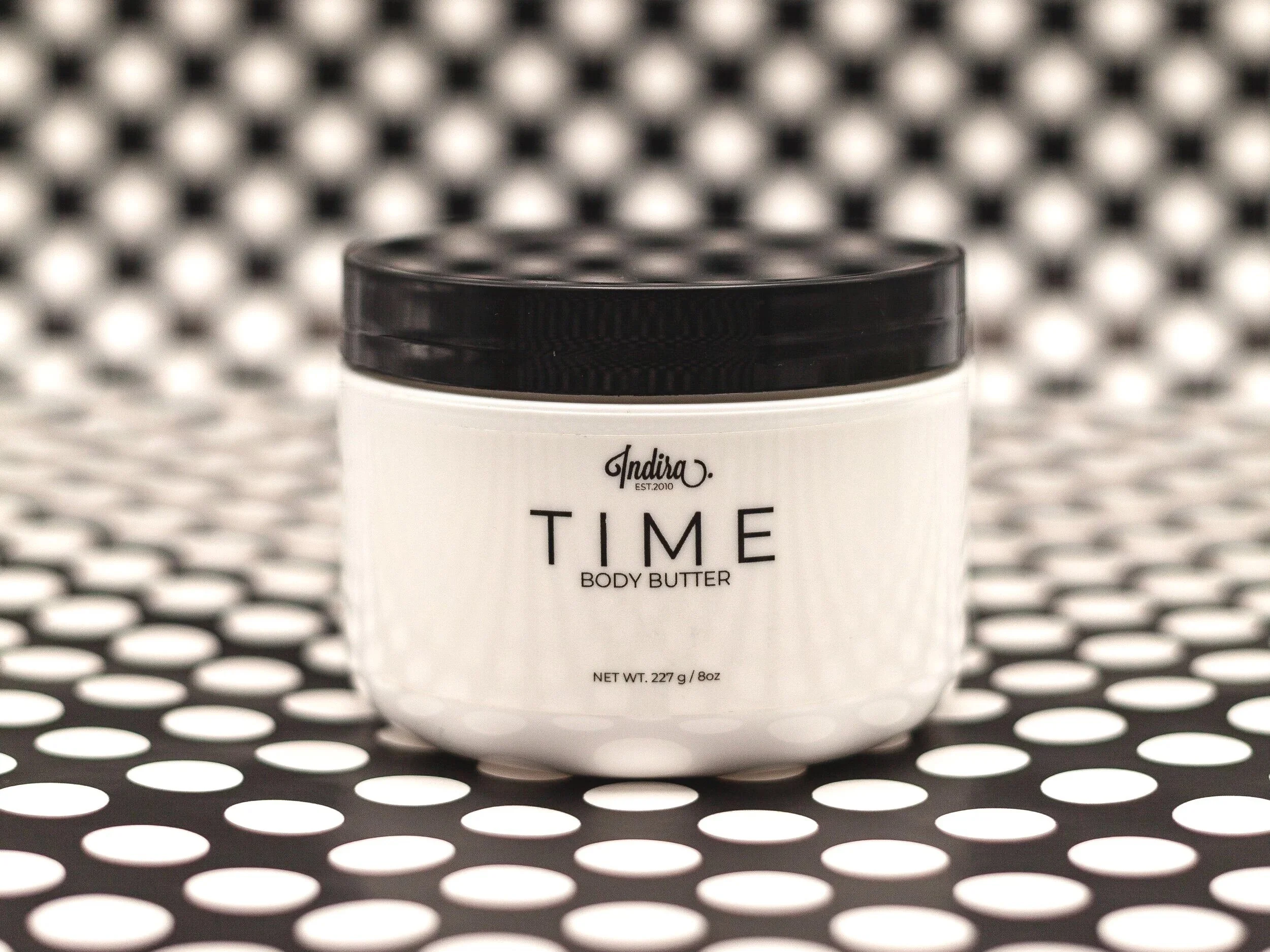 A white container of body butter with a black lid, labeled 'Indira Time Body Butter', placed on a black and white polka dot surface with a polka dot background.
