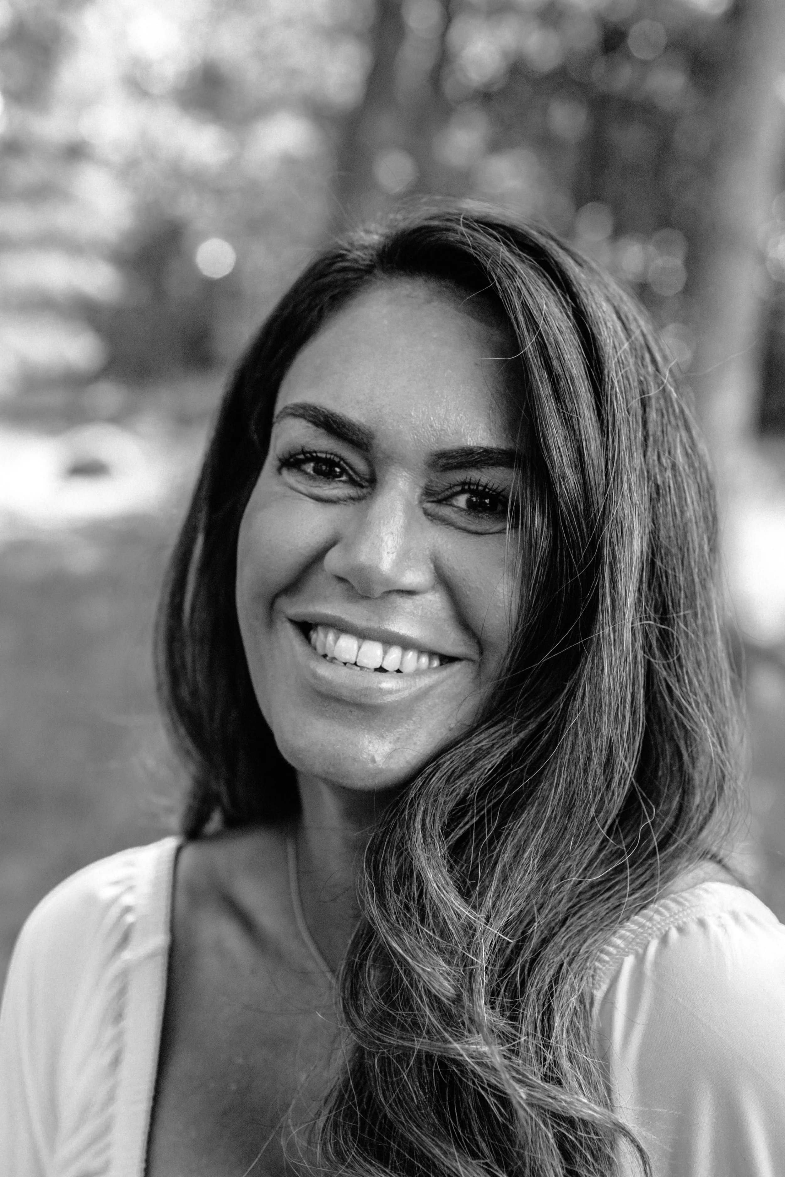 Svartvitt porträtt av en kvinna som ler, med långt, vågigt hår, utomhus med suddigt bakgrund av träd.