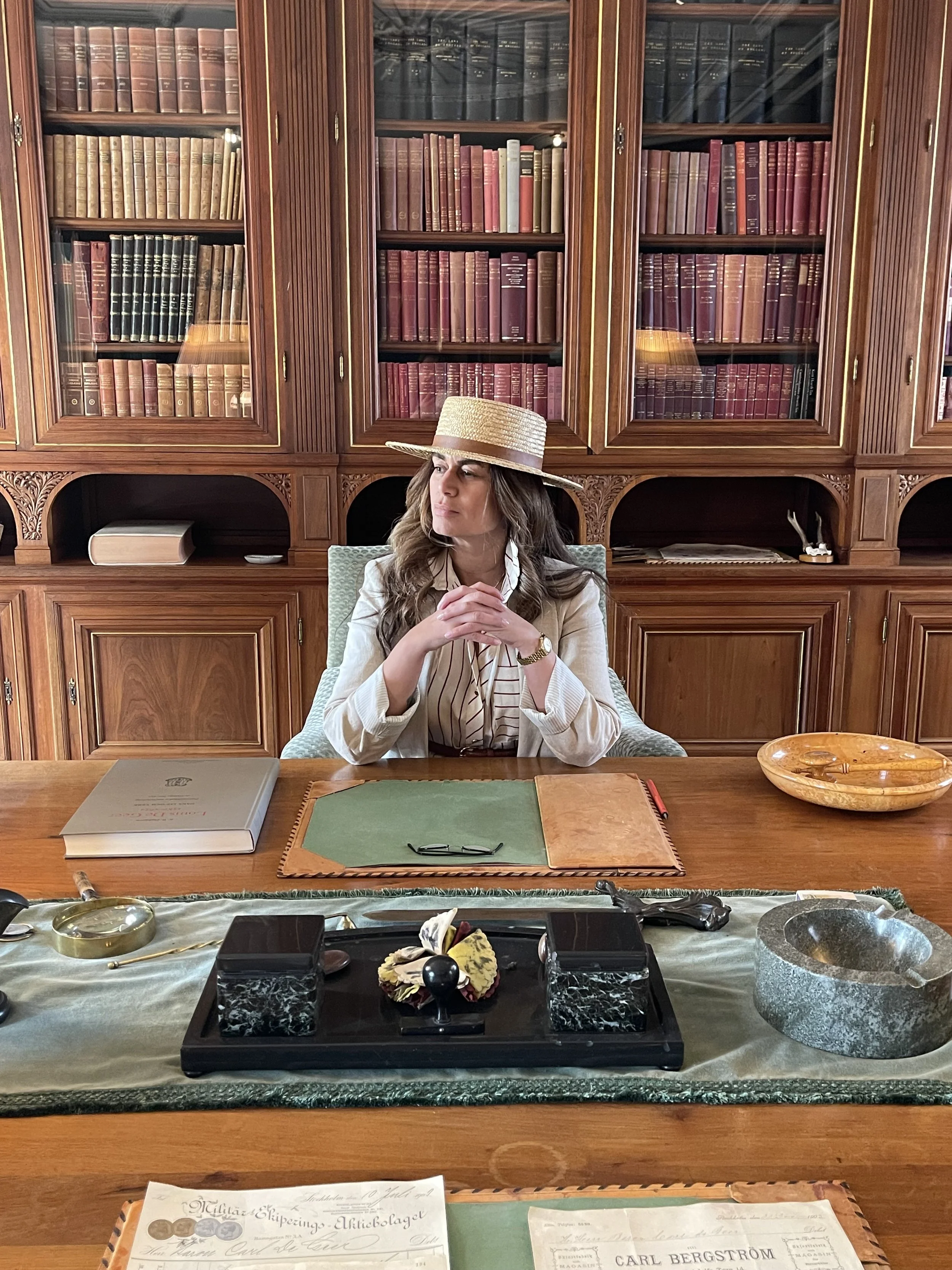 En kvinna med pratmössa sitter vid ett stort skrivbord i ett rum med biblioteksväggar fyllda med böcker.