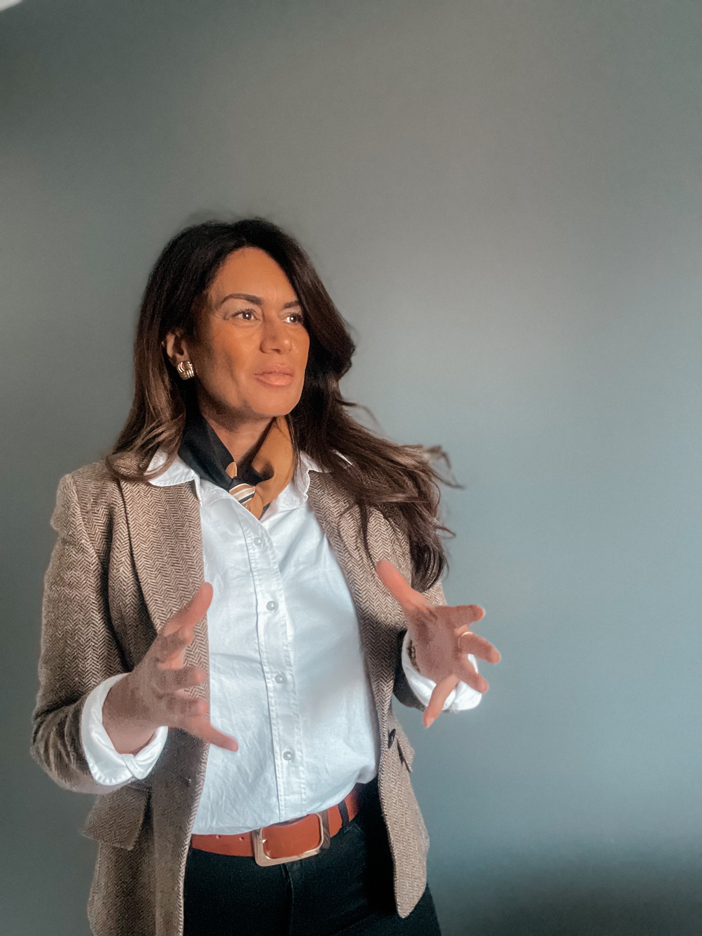 Kvinna som pratar eller förklarar, iförd vit blus, beige kavaj och svart scarf, står mot en neutral bakgrund.