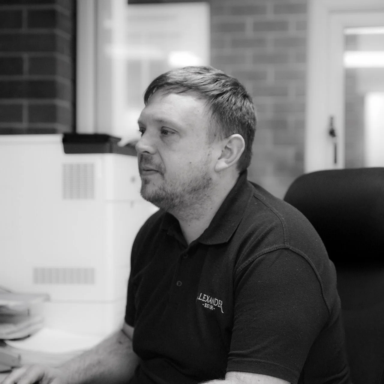Black and white photo of a man sitting at a desk in an office environment.